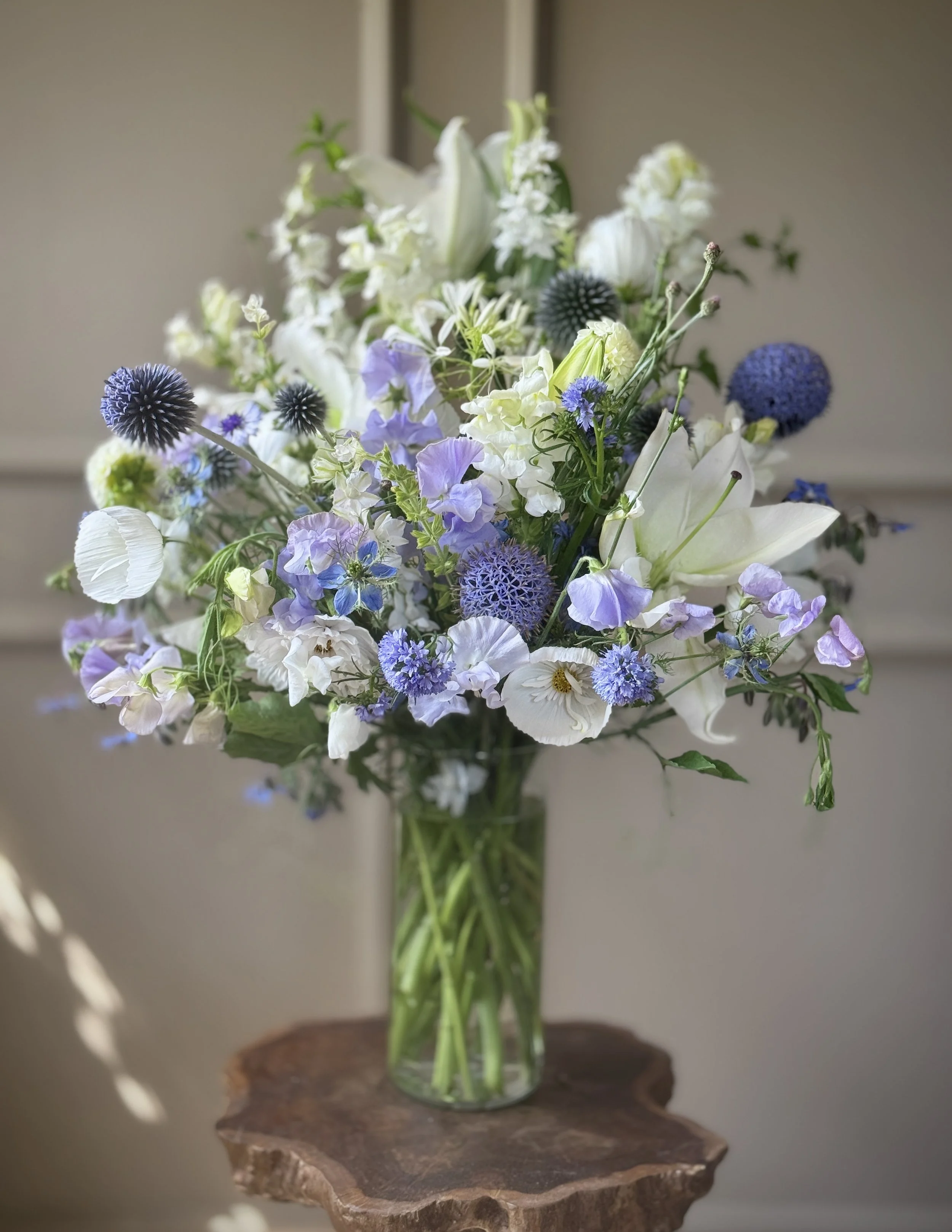 A clear glass vase filled with a bouquet of white, purple, and green flowers, placed on a wooden surface against a light background.