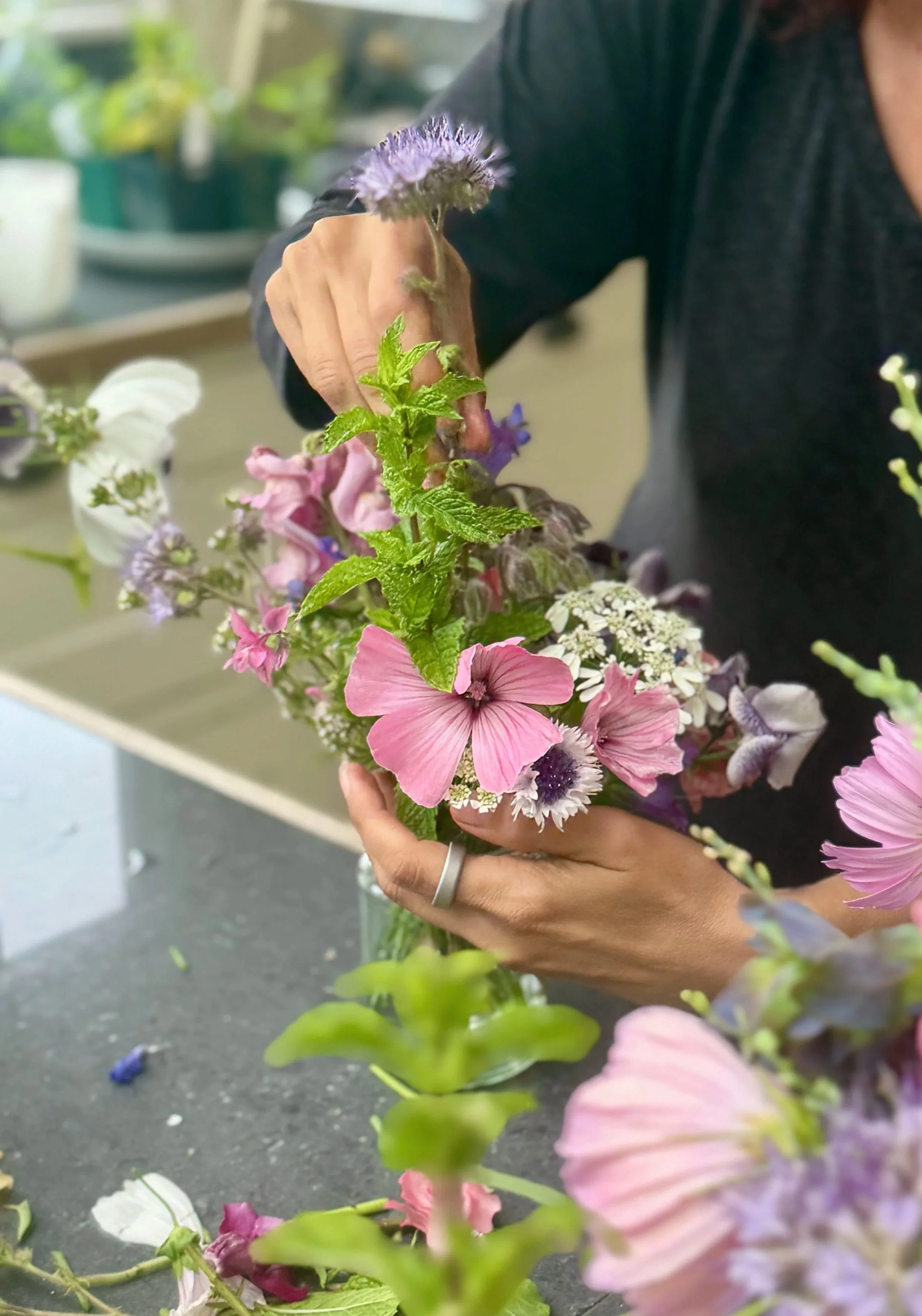 Person arranging colorful flowers, including pink, purple, and white blooms, on a dark surface in a well-lit room.