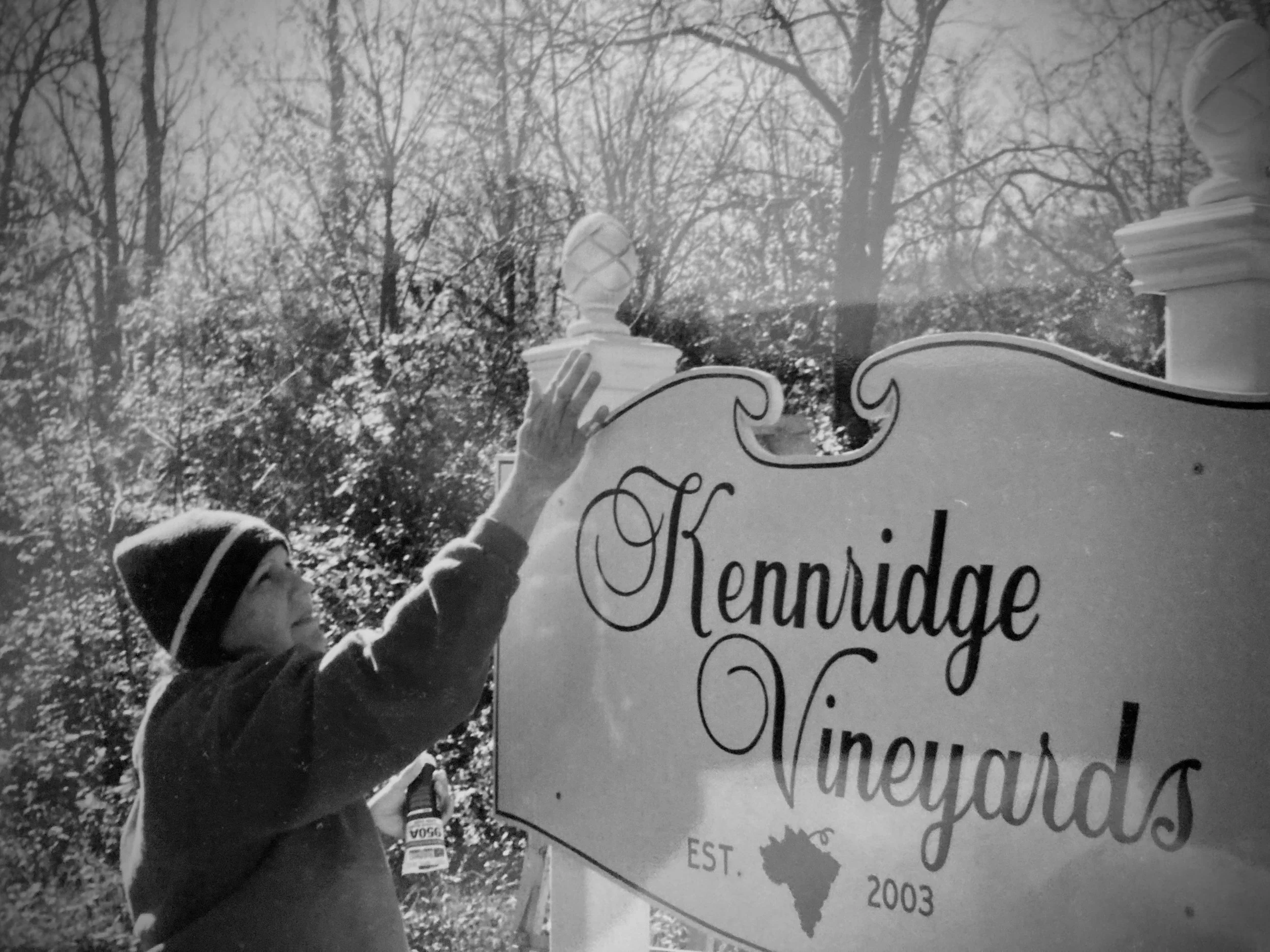 Child holding a bottle next to a sign with decorative cursive text, outdoors with leafless trees in the background.