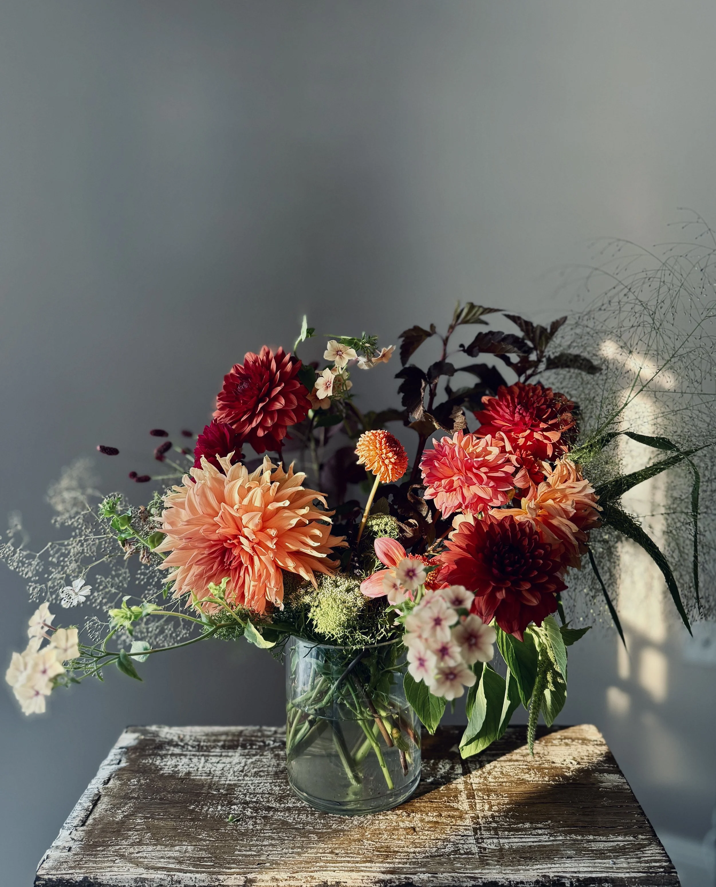 A bouquet of mixed flowers in a glass vase on a rustic wooden table, against a neutral wall background.