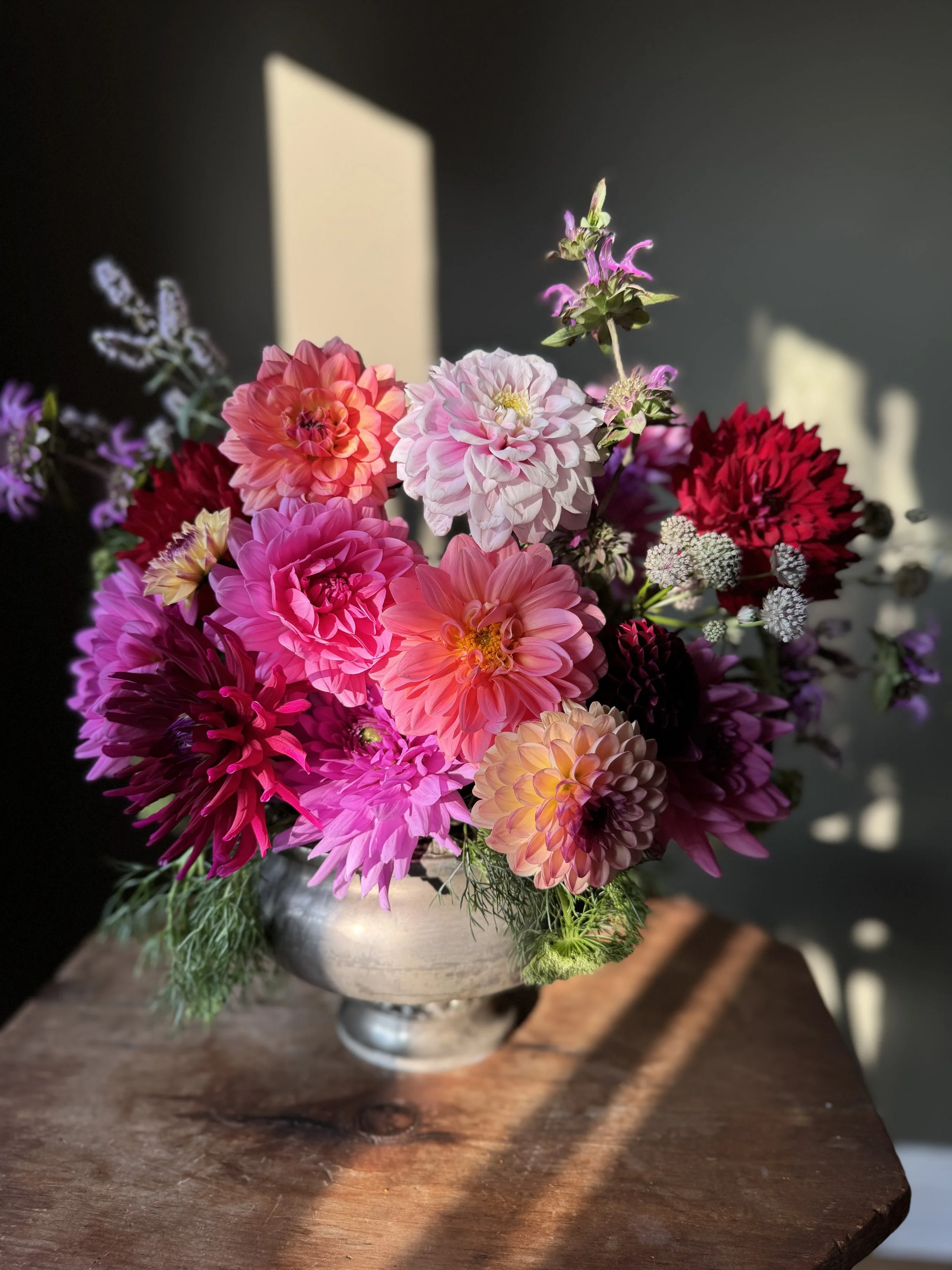 A bouquet of pink, purple, red, and white flowers in a metal vase on a wooden table, with sunlight casting shadows on a dark wall.
