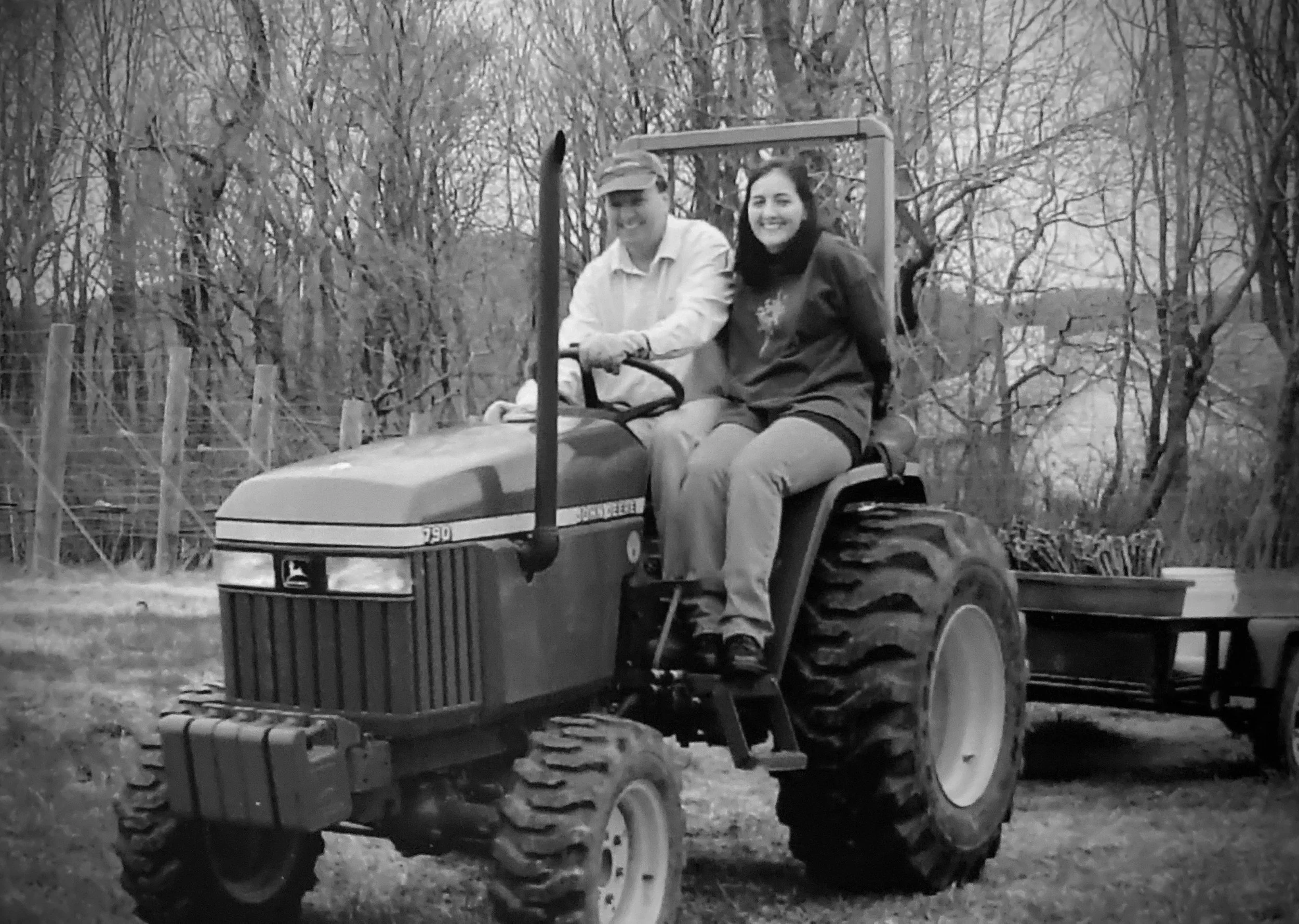 A black and white photo of a man and woman sitting on a small tractor outdoors, surrounded by leafless trees. The man is wearing a hat and light-colored shirt, and the woman is wearing a dark jacket and has long dark hair.