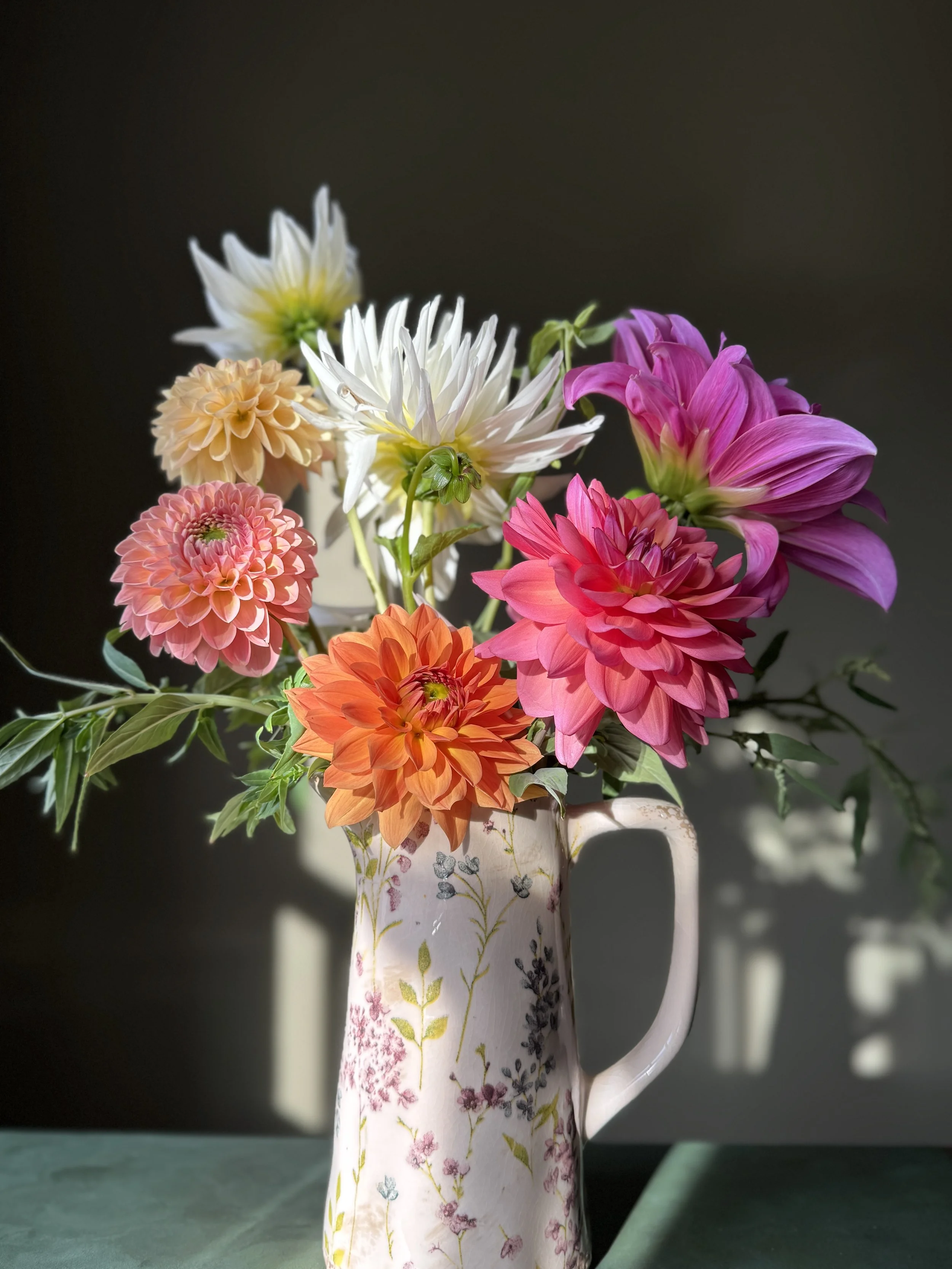 A ceramic pitcher with floral patterns filled with a variety of colorful flowers including pink, orange, white, and purple dahlias and white chrysanthemums, placed on a green surface with a dark background.