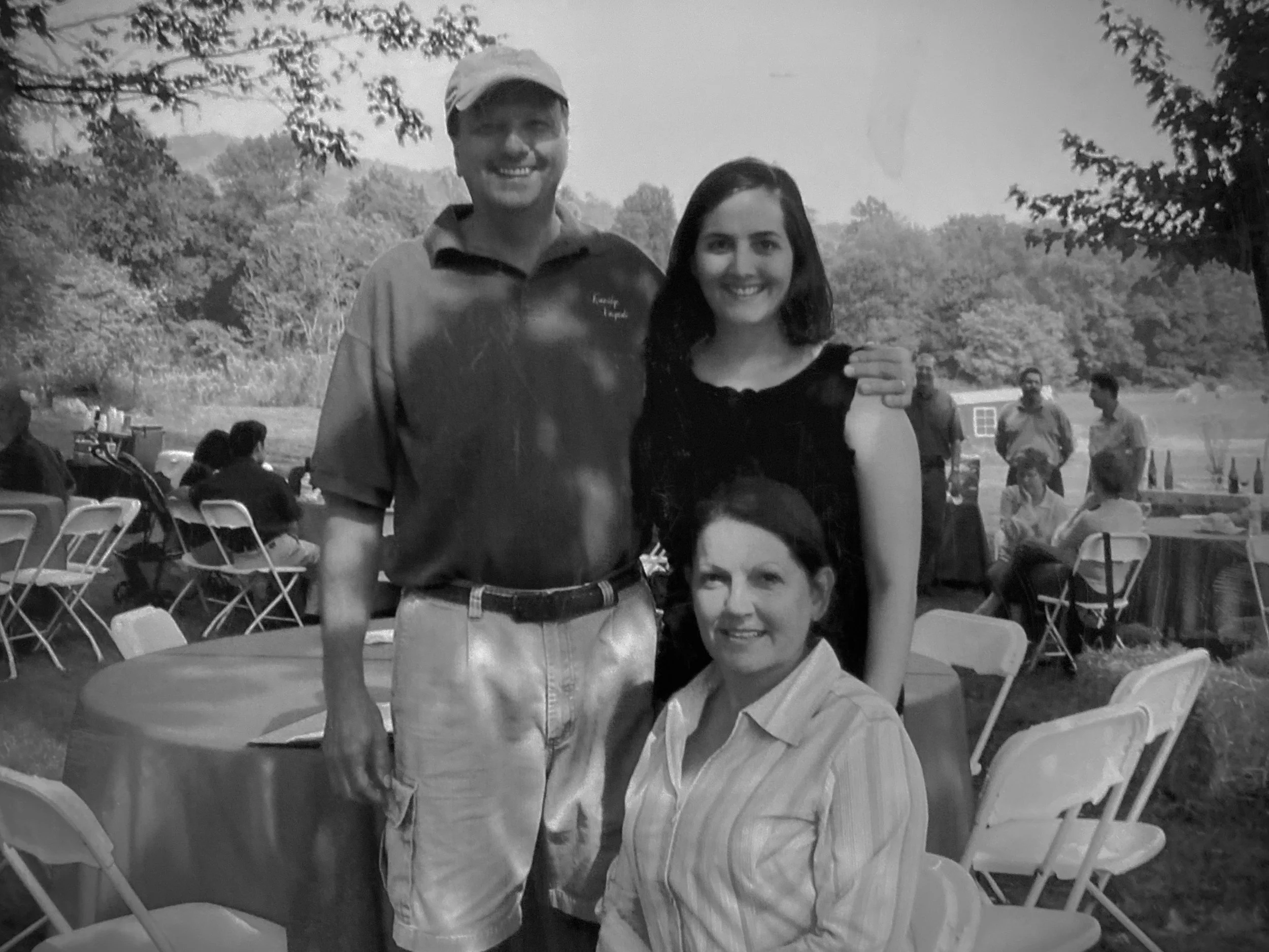 A black and white photo of two smiling people at an outdoor gathering. One man with a beard and cap is standing behind a woman with dark hair, who is sitting. They are surrounded by other people and chairs, with trees and a hilly landscape in the background.