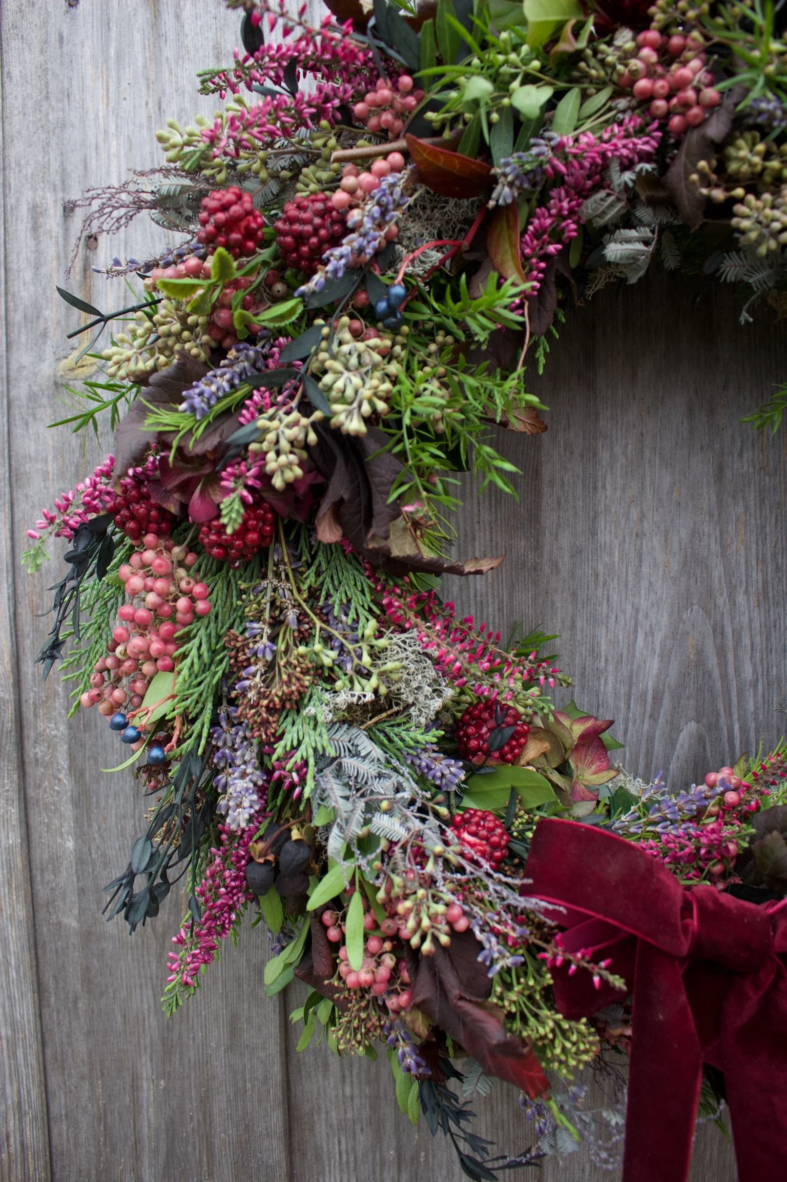 A holiday wreath decorated with pink, purple, and red berries, green leaves, and a burgundy velvet ribbon, hanging on a weathered wooden surface.
