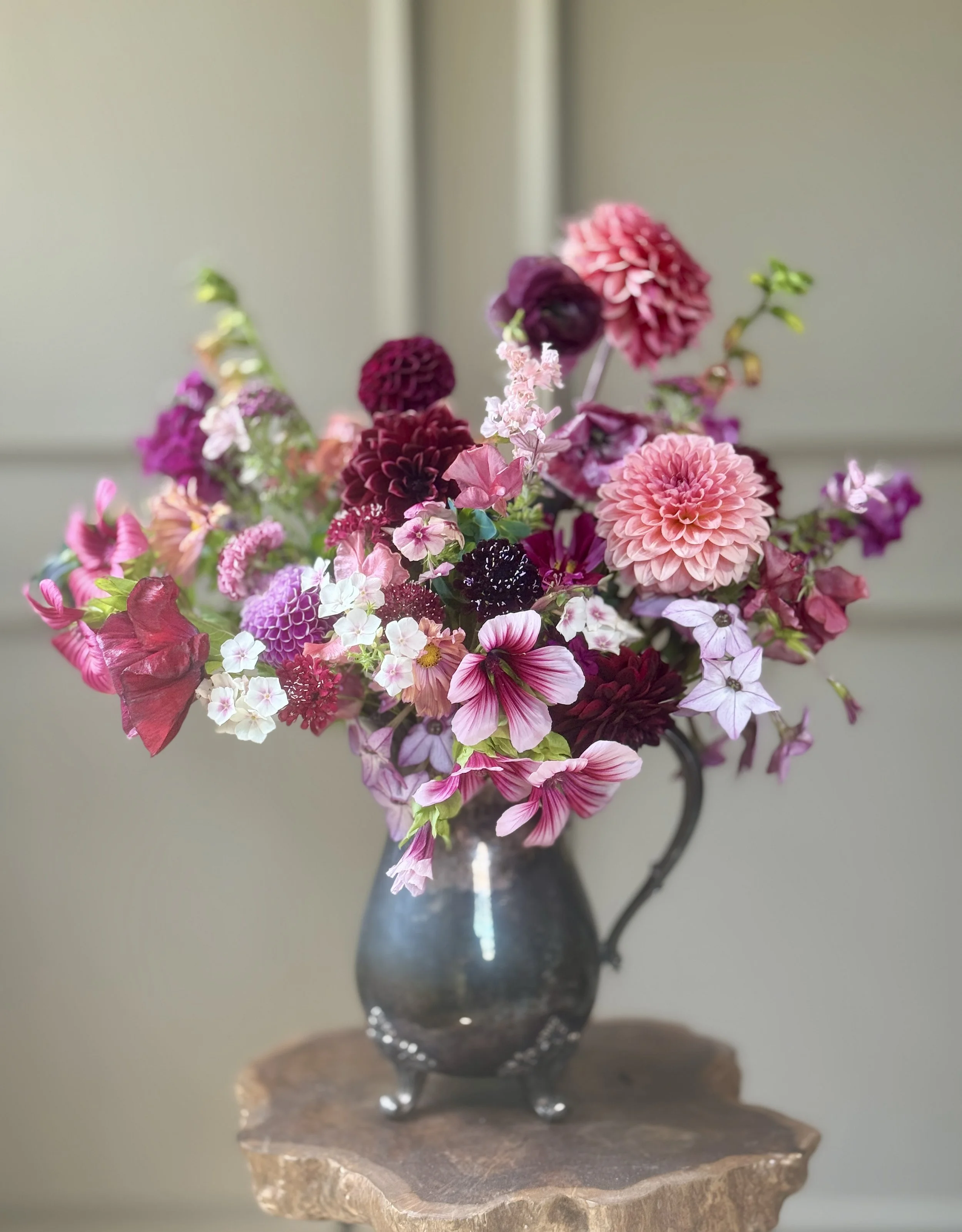 A colorful bouquet of various pink, purple, and white flowers in a dark, reflective vase on a rustic wooden stand.