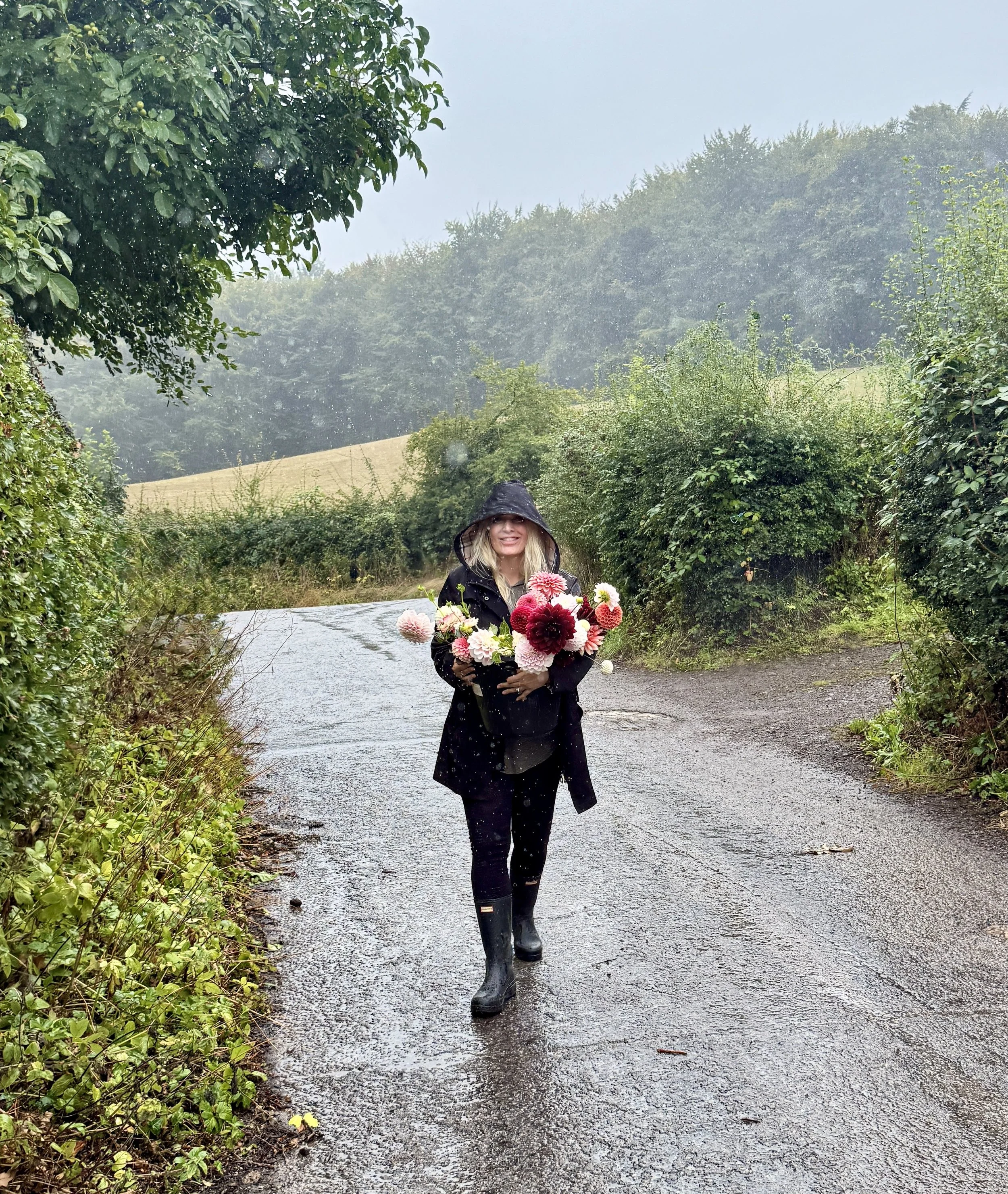 A woman walking on a rain-soaked country road holding a bouquet of pink, white, and red flowers, wearing black rain boots, a black jacket, and a hood, with lush green trees and hills in the background.