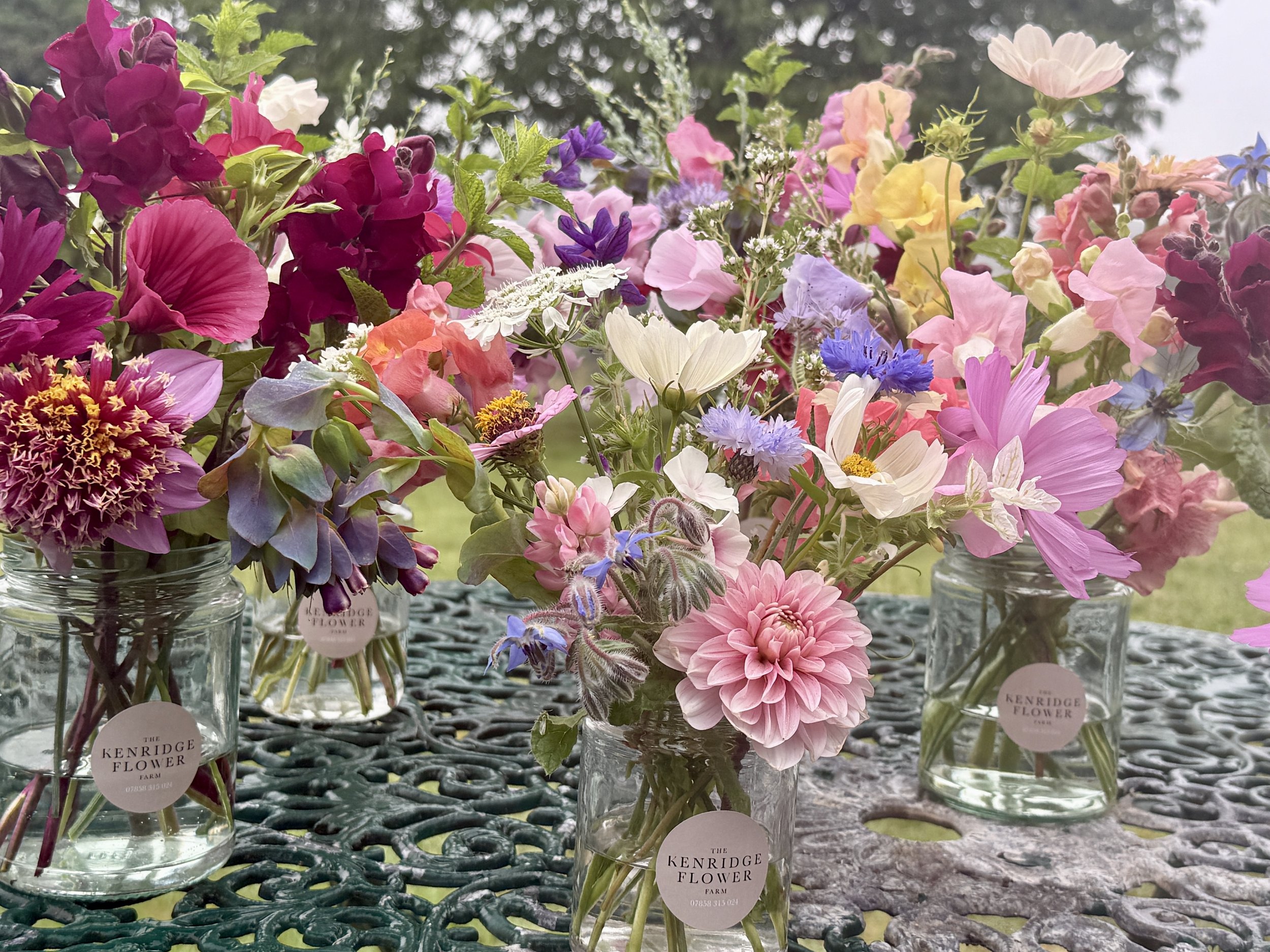 A colorful assortment of flowers in glass jars on a table with a outdoor setting and trees in background.