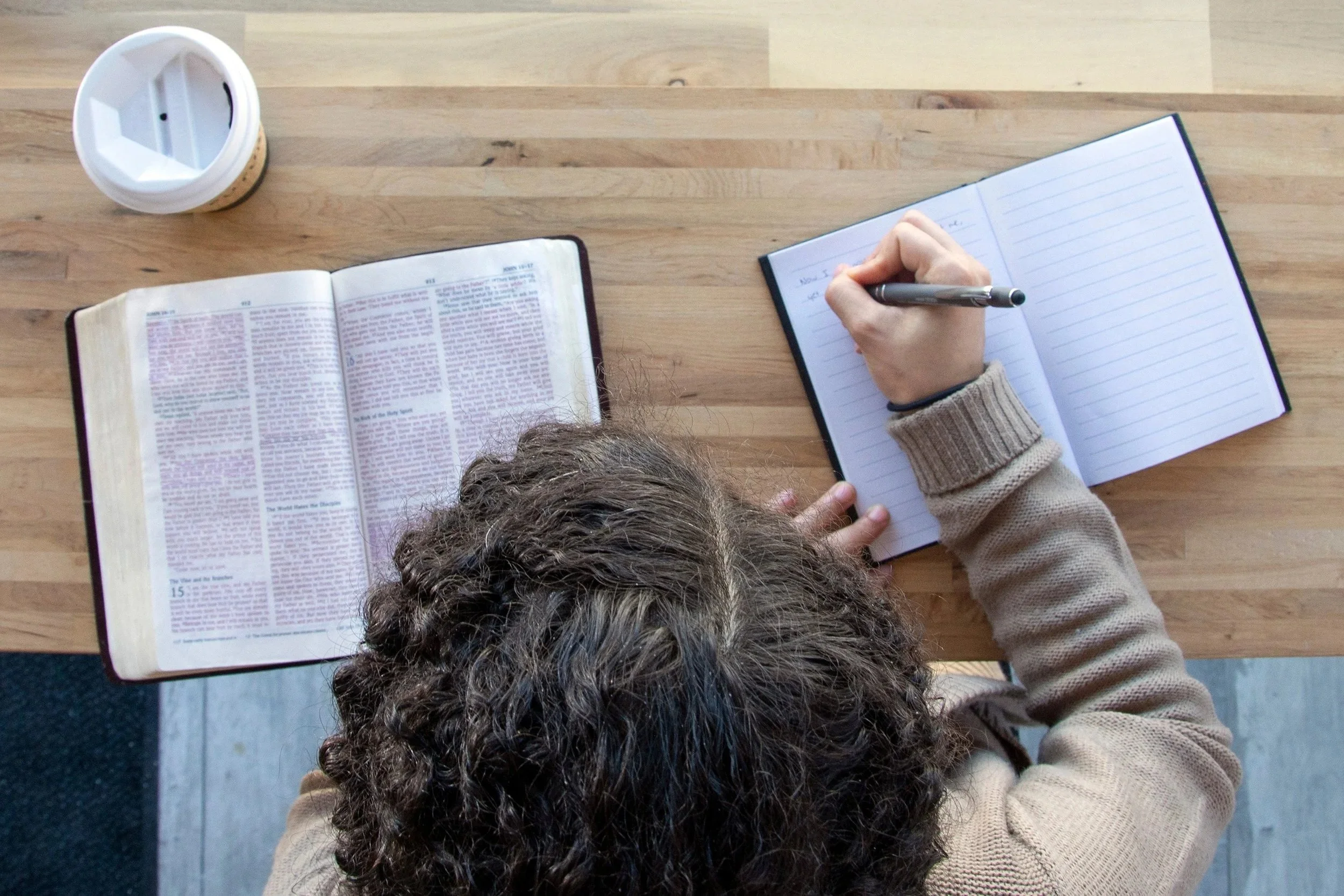 A person with curly hair sitting at a wooden table, writing in a notebook with a pen, with an open Bible and a coffee cup on the table.