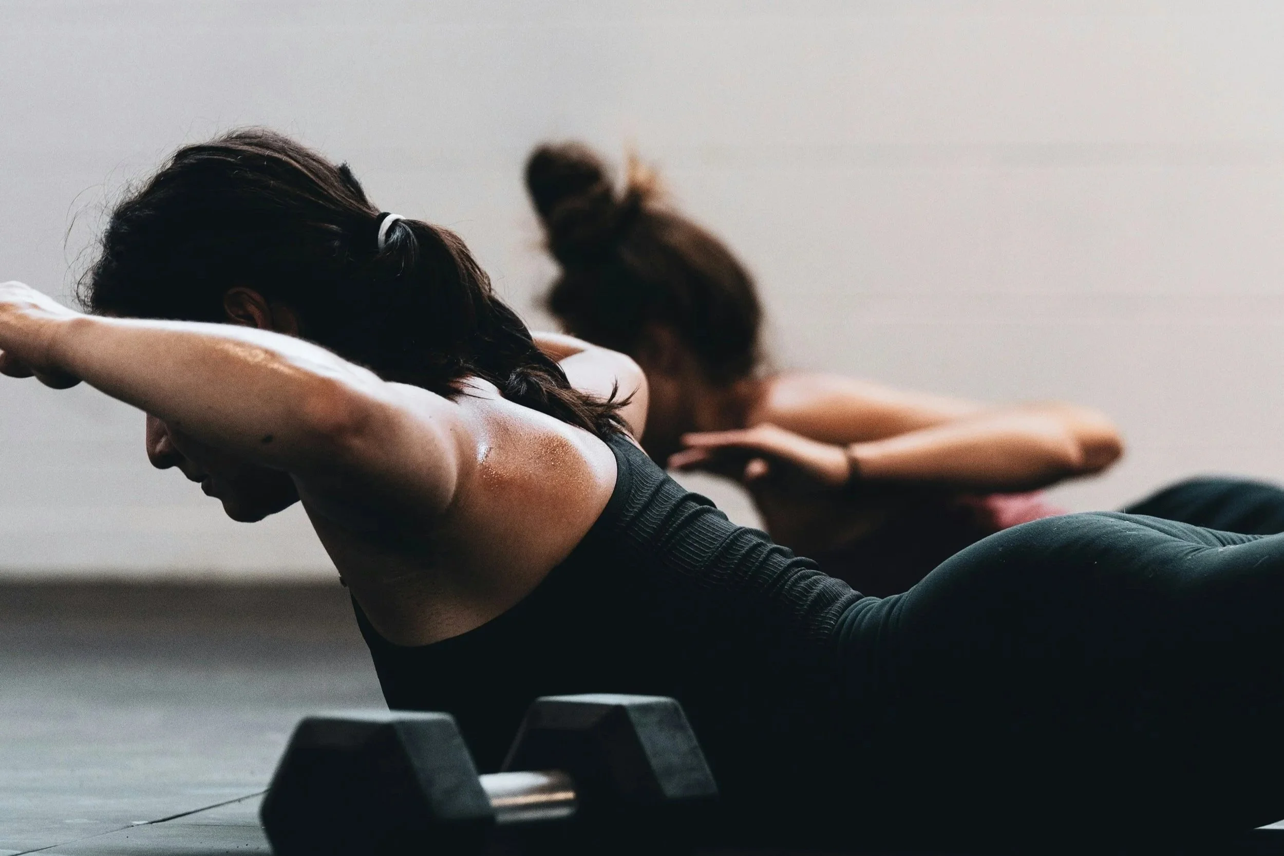 Two women doing a core workout on the floor, with one woman in the foreground and another in the background, both in a gym setting.