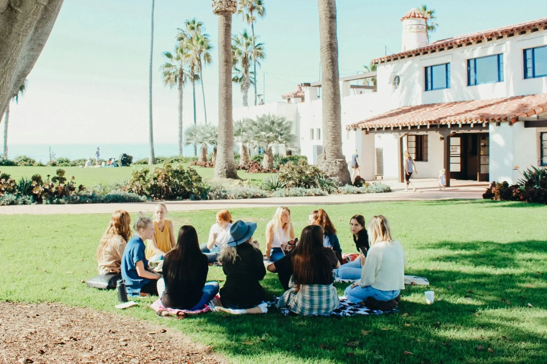A group of women sitting in a circle on a blanket on the grass in a park near a white building with a red-tiled roof, surrounded by palm trees, on a sunny day with clear blue sky.