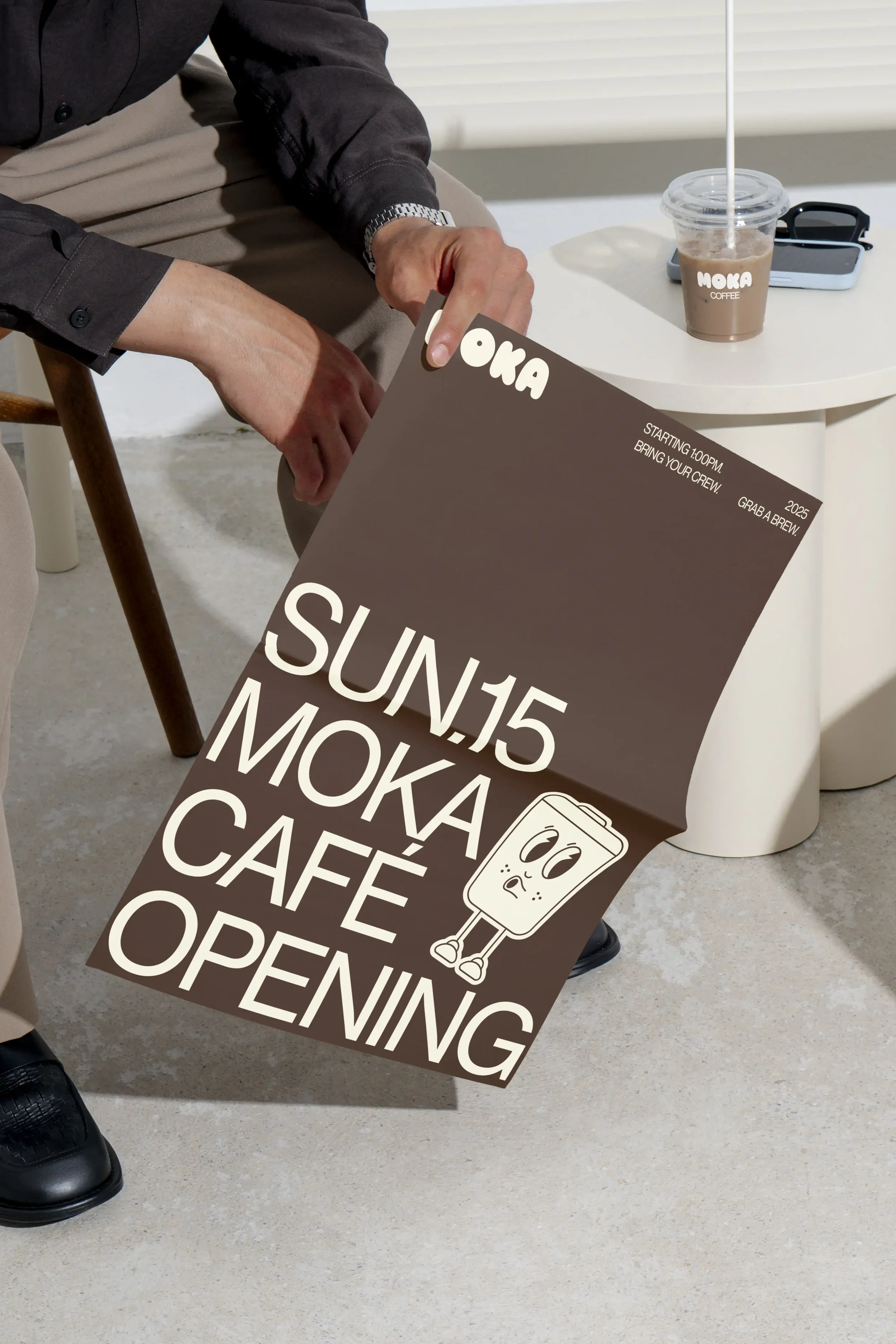 A person holding a Moka coffee carrier with two cups, sitting on a bench. The carrier has a cartoon coffee phone illustration and the words 'Dark Roast Blend' and 'Moka' on it.