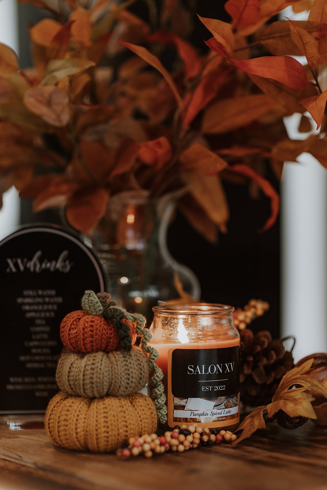 A cozy autumnal display featuring a lit scented candle labeled 'SALON XV Pumpkin Spiced Latte', a small knitted pumpkin stacked with a crocheted orange pumpkin, a vase with orange leaves on a wooden surface.