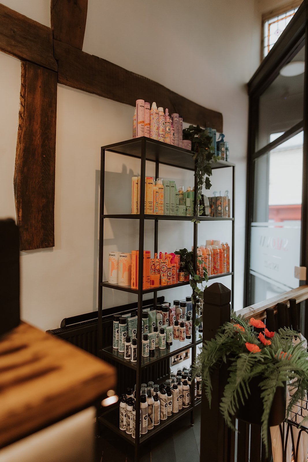 A black metal shelving unit with various hair products, some labeled, placed in a welcoming indoor space with a large window and a potted fern with red flowers nearby.