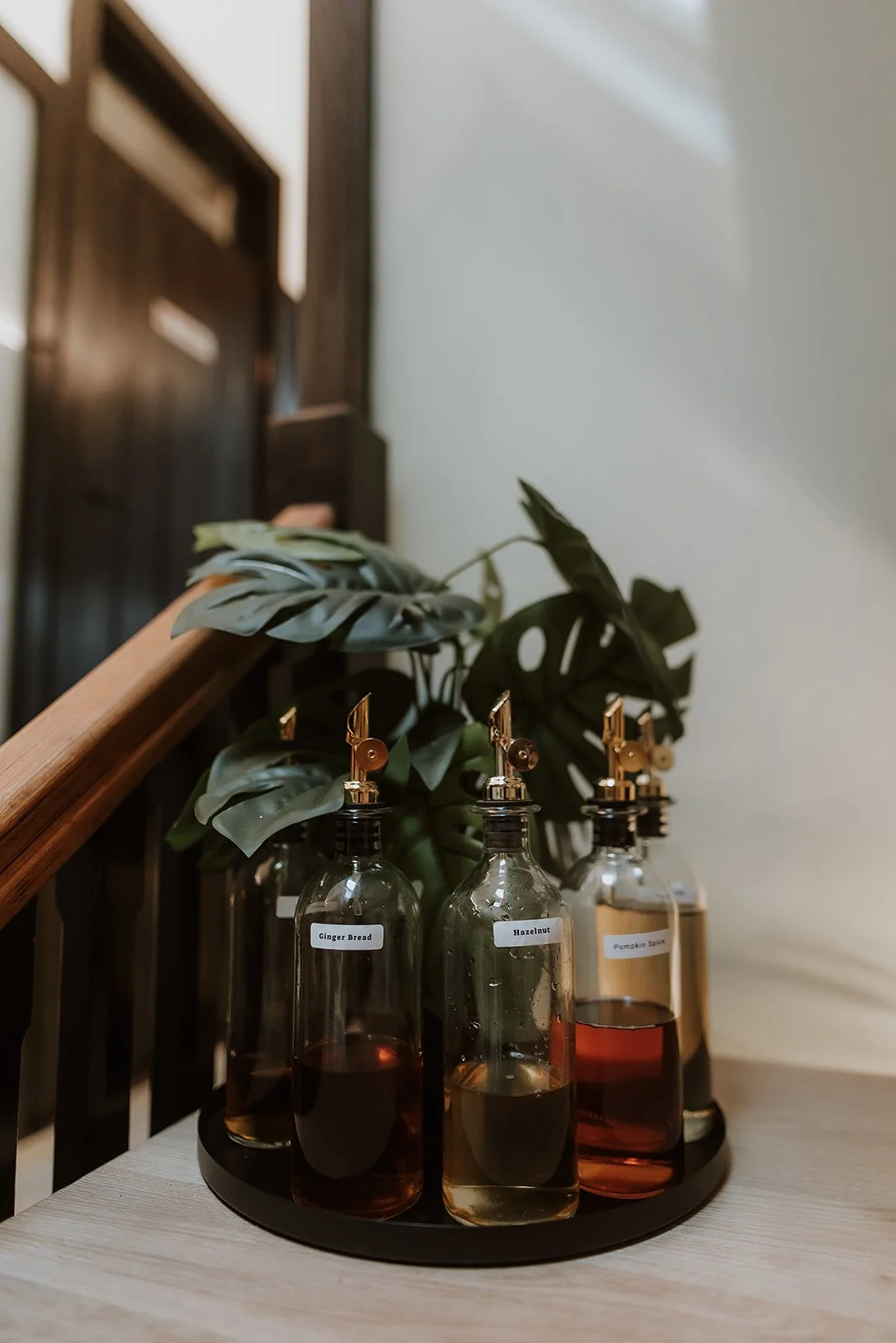 Set of coffee syrups labelled Ginger Bread, Hazelnut, Pumpkin Spice, on a round tray with a plant and wooden staircase nearby.