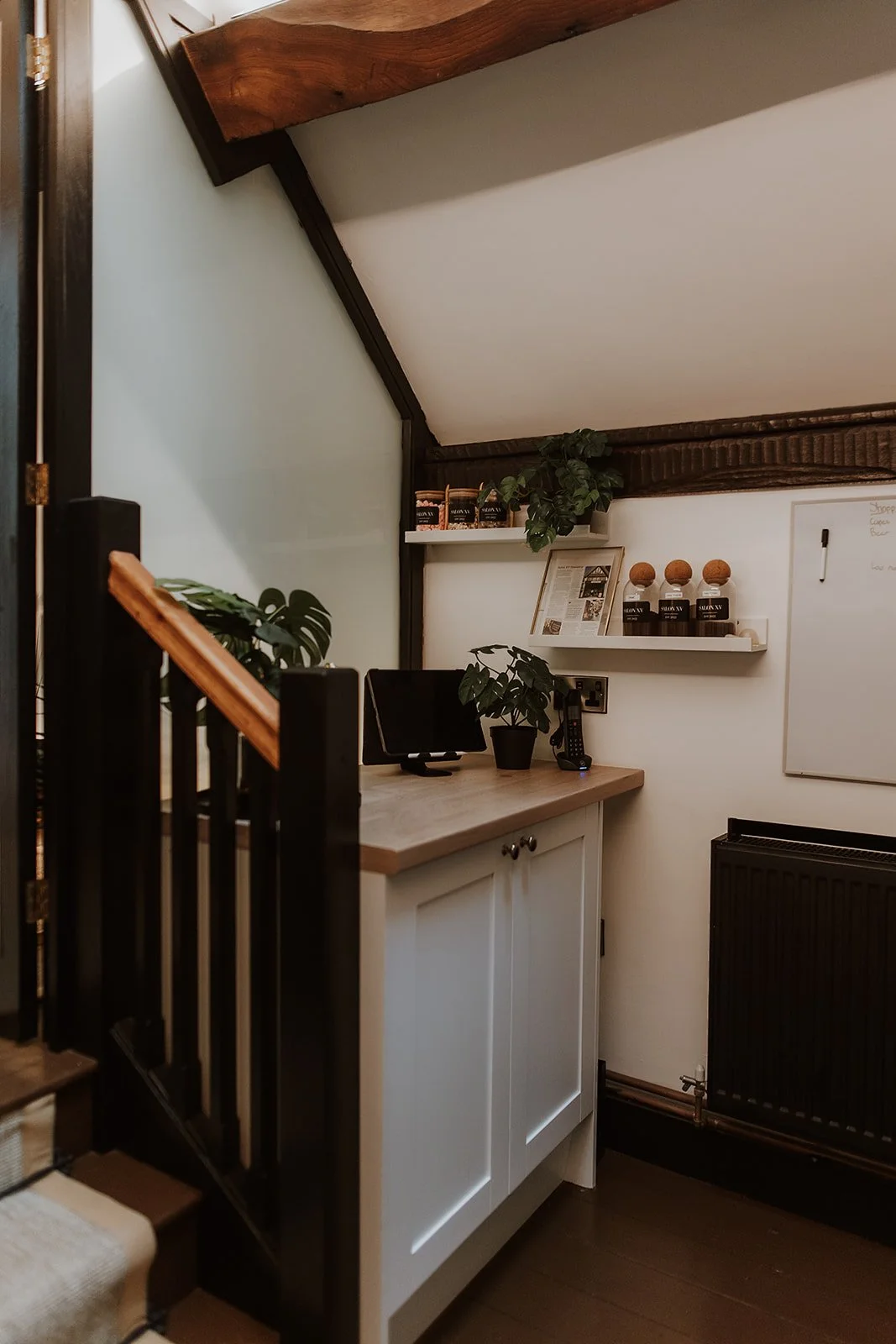 Corner of the kitchen at Salon XV, a wooden countertop, potted plants, a small monitor, a telephone, shelves with jars of coffee and decorative items, and a whiteboard on the wall.