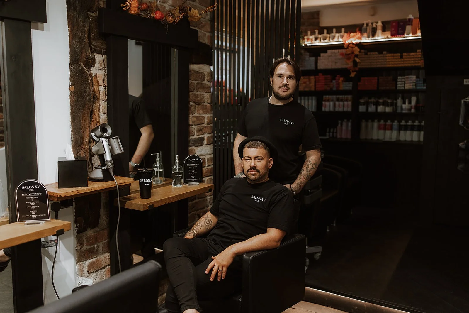 Sam & Martyn Lewis-Evans, Salon XV co-founders, pose for a photo in a salon chair, wearing black Salon XV branded shirts. The salon interior features a brick wall, a mirror, and hair products on shelves.