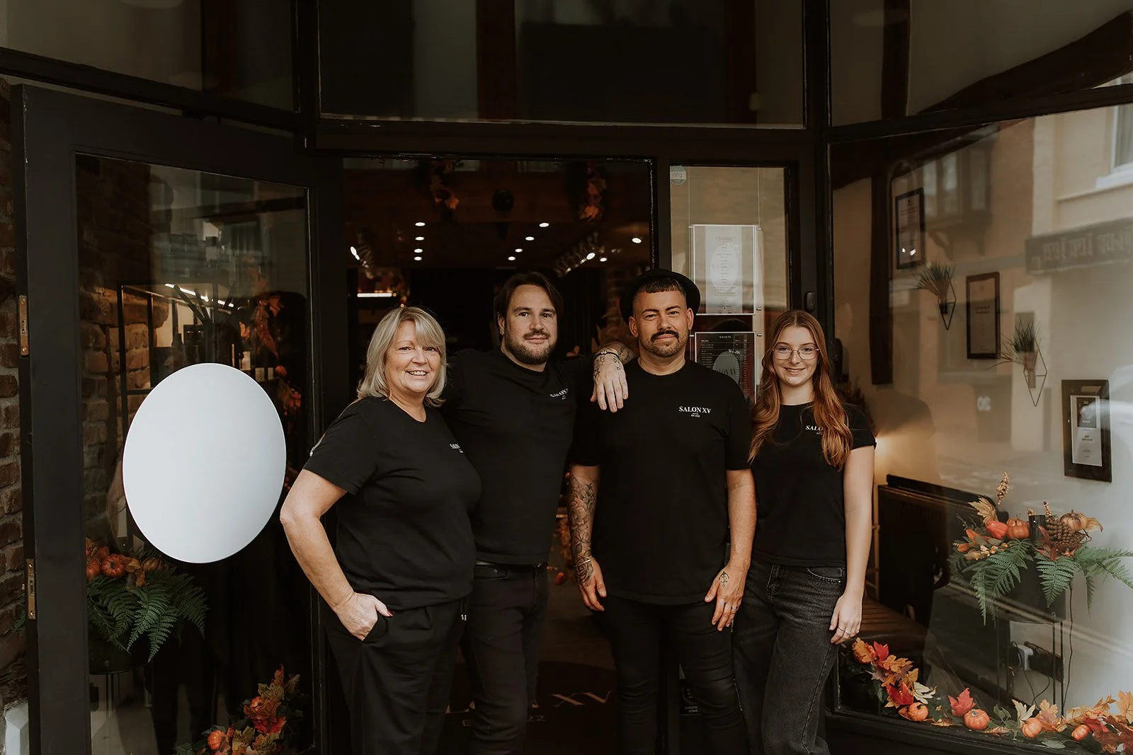 Debbie, Martyn Lewis-Evans, Sam Lewis-Evans & Abbie Williams standing at the entrance of Salon XV, dressed in branded black T-shirts, smiling for a photo.