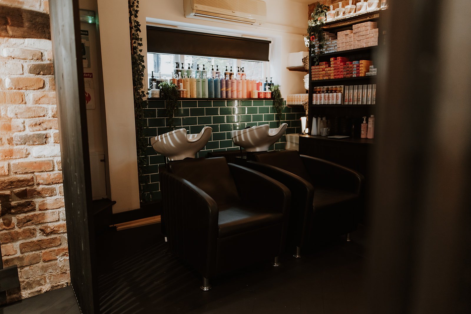 Two black salon chairs with white shampoo sinks behind them, inside Salon XV in Oswestry with brick and green tile walls and shelves stocked with hair products.