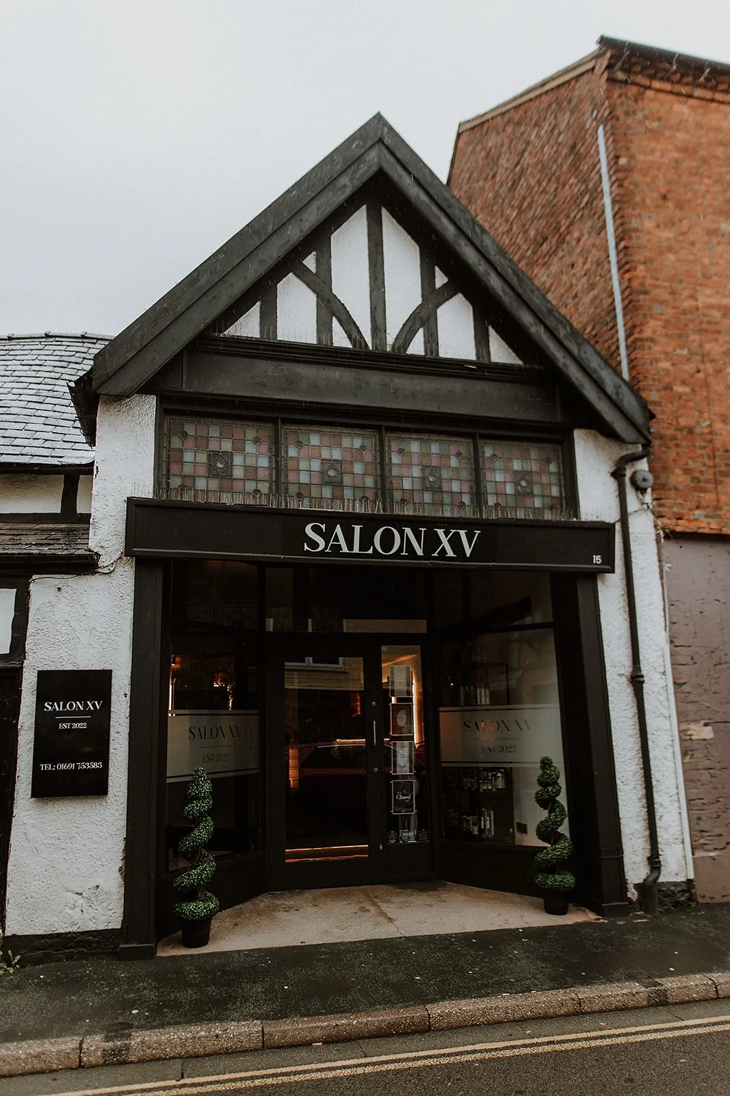 Exterior of a building on Beatrice Street in Oswestry with a sign saying 'Salon XV,' featuring black framing, stained glass windows, two potted spiral topiary plants, and a brick building beside it.