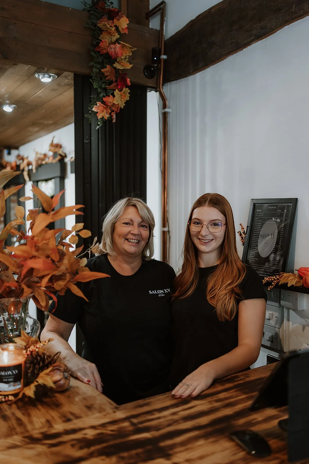 Debbie & Abbie Williams standing behind a wooden Salon counter, smiling, with autumn leaf decorations on the wall and counter.