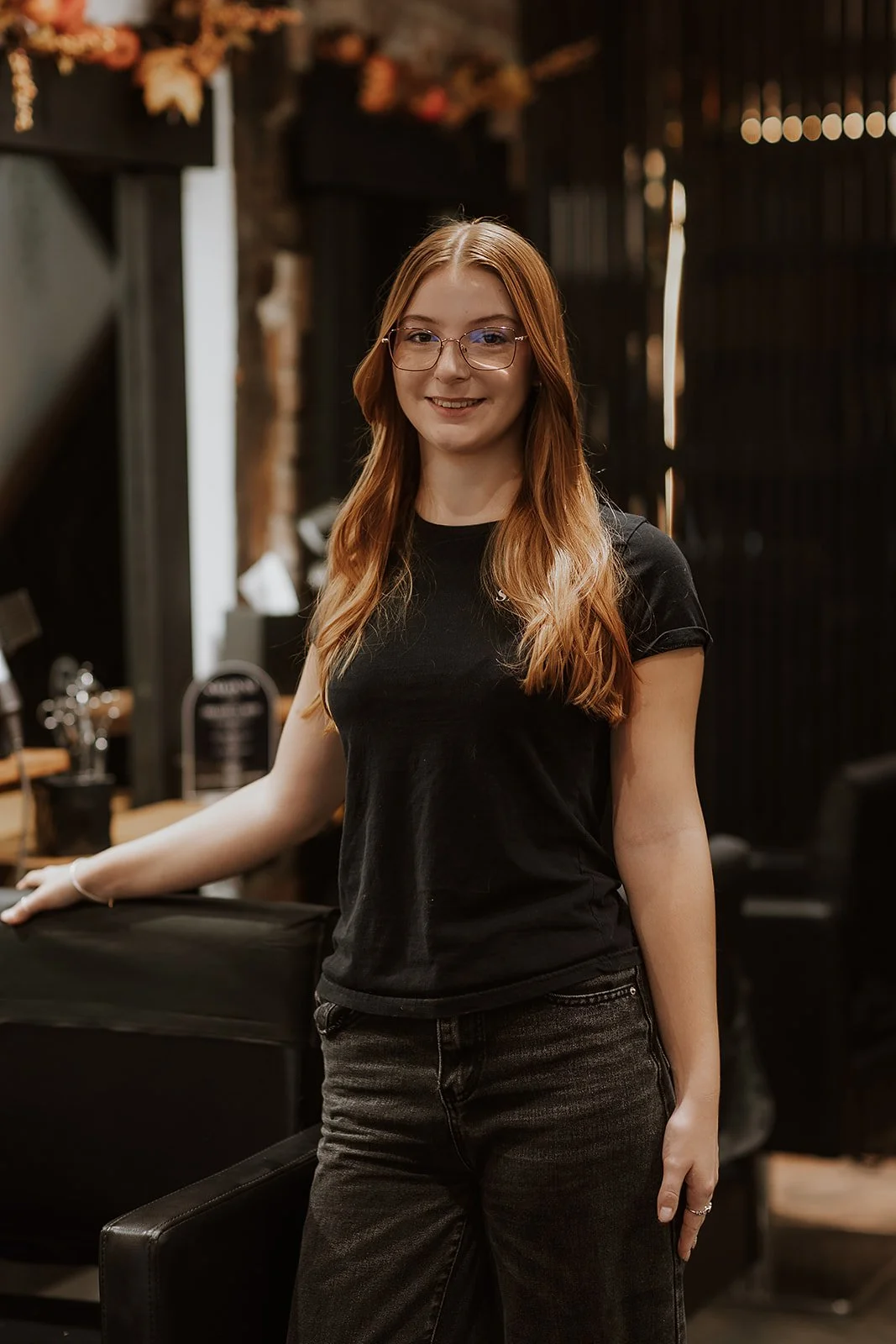 Abbie Williams, Junior Stylist at Salon XV. A young woman with long red hair, wearing glasses and a black t-shirt, is standing indoors with a smile.