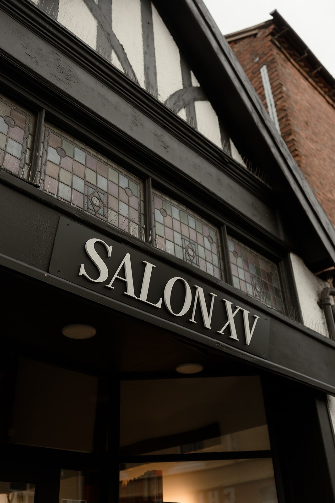 Close-up of a black sign that reads 'SALON XV' above the entrance of a building with decorative stained glass windows and timber framing.