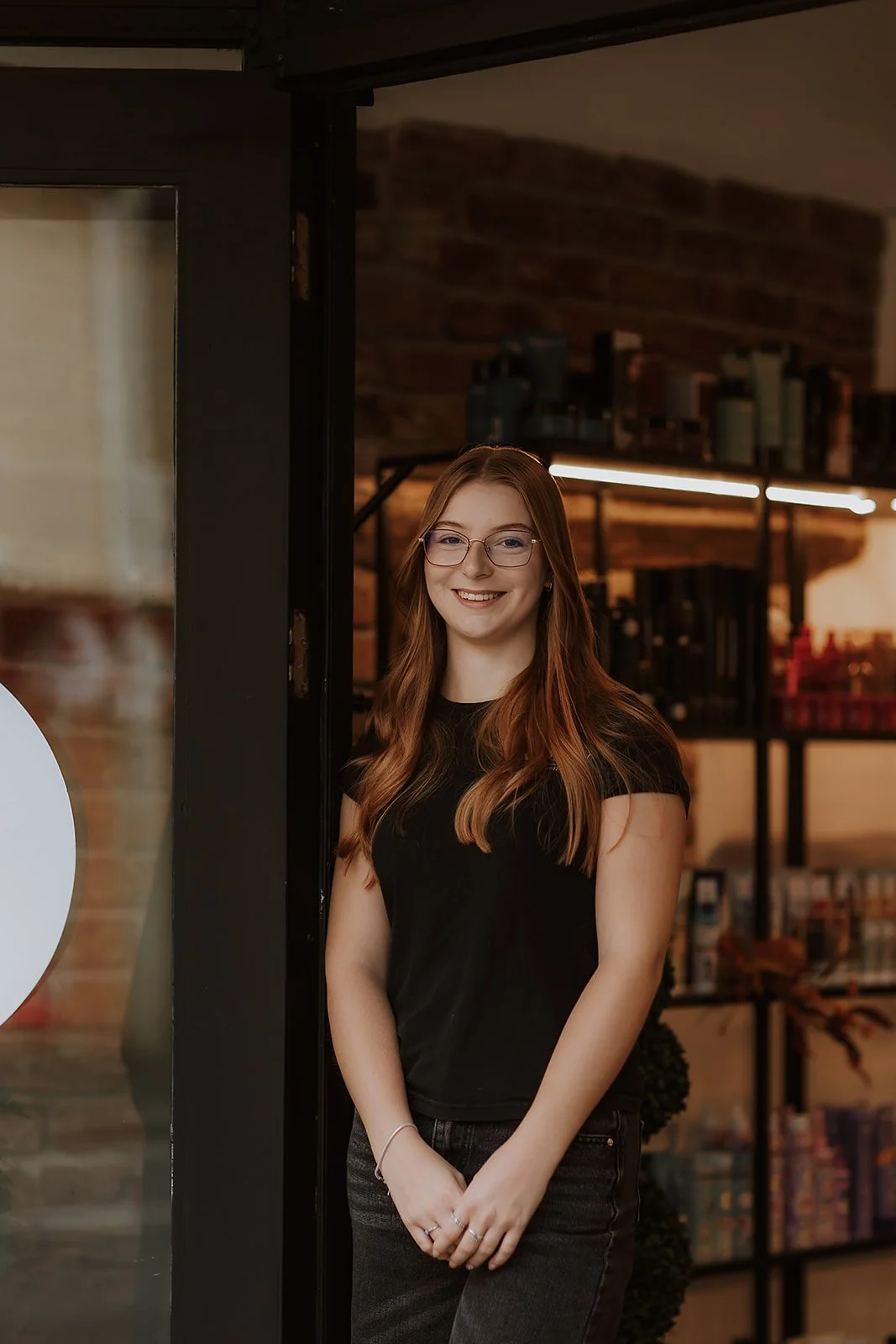 Abbie Williams, with long red hair and glasses, wearing a black t-shirt and dark jeans, is smiling and standing outside Salon XV, with wooden and brick decor.