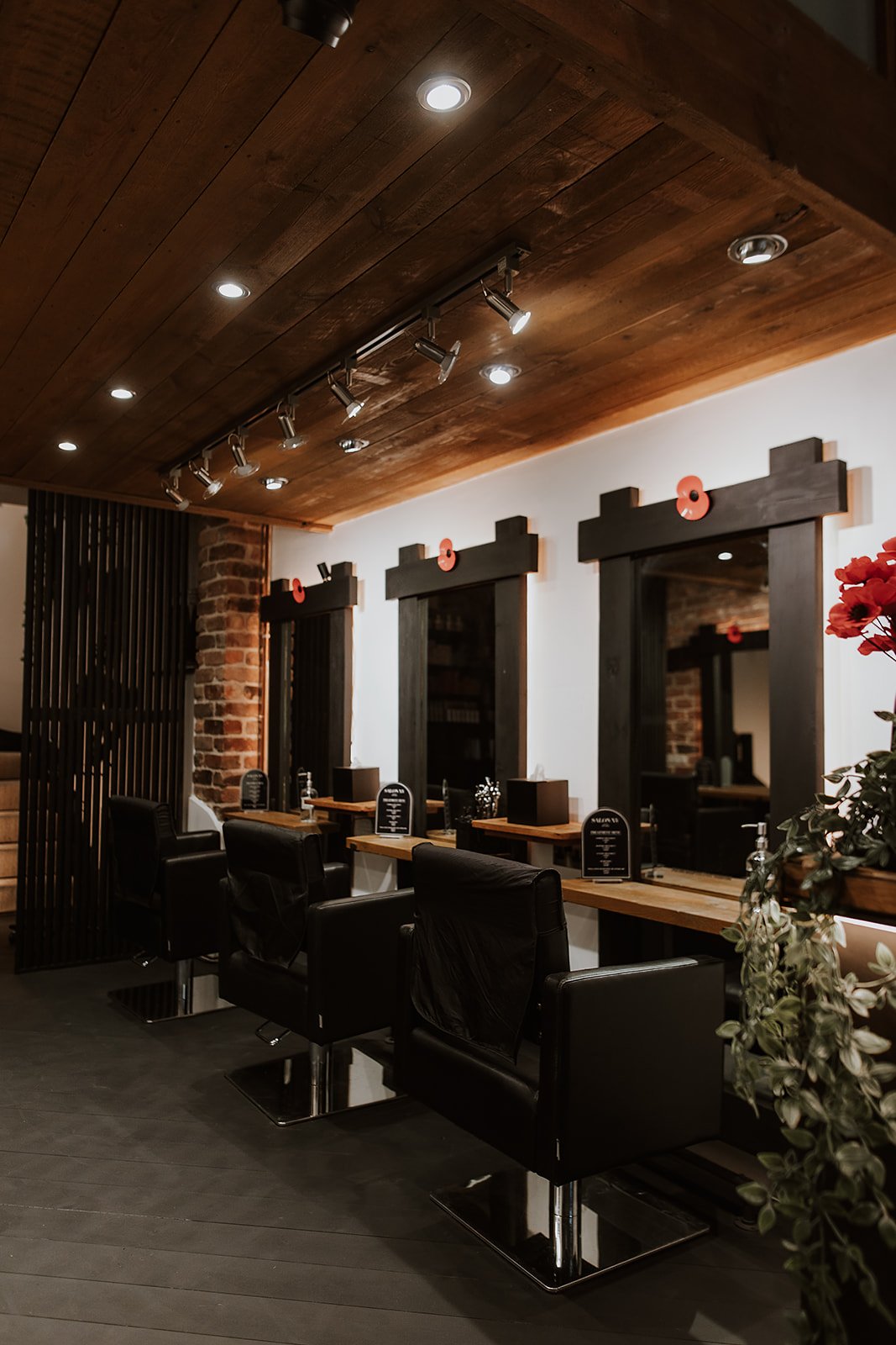 Interior of Salon XV in Oswestry with three styling chairs facing mirrors on a white wall, with a wooden ceiling, exposed brick pillars, and black wooden frames around the mirrors. There are small tables, and a decorative poppies.