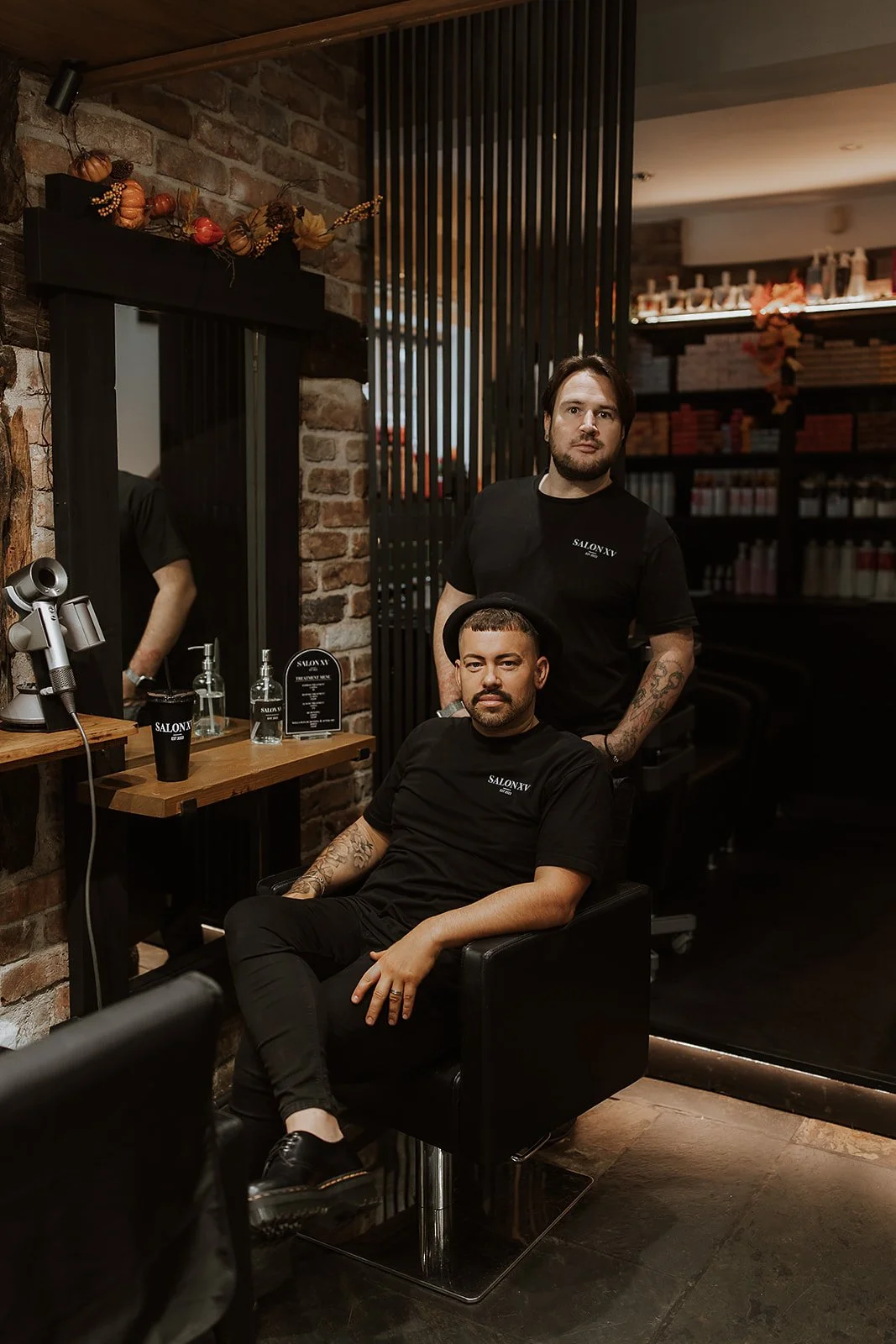 Sam & Martyn Lewis-Evans, Salon XV co-founders, pose for a photo in a salon chair, wearing black Salon XV branded shirts. The salon interior features a brick wall, a mirror, and hair products on shelves.