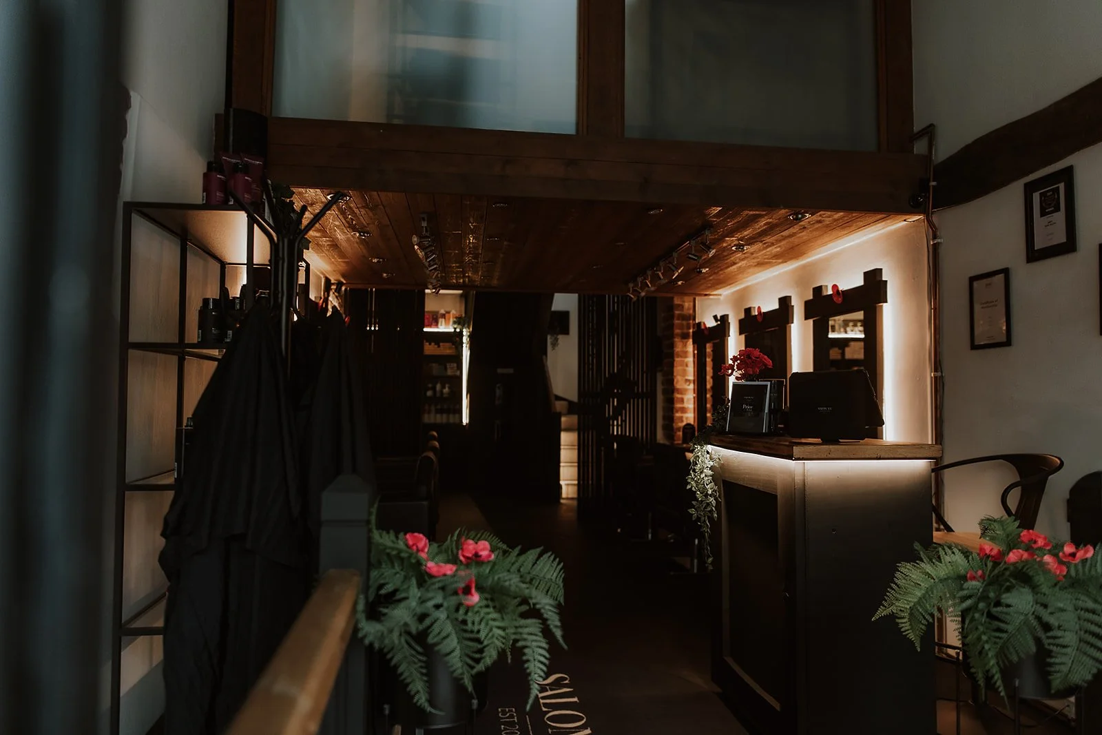 Interior of Salon XV in Oswestry, with a wooden ceiling, warm lighting, a counter with black and white decor, and small plants with red flowers near the entrance.