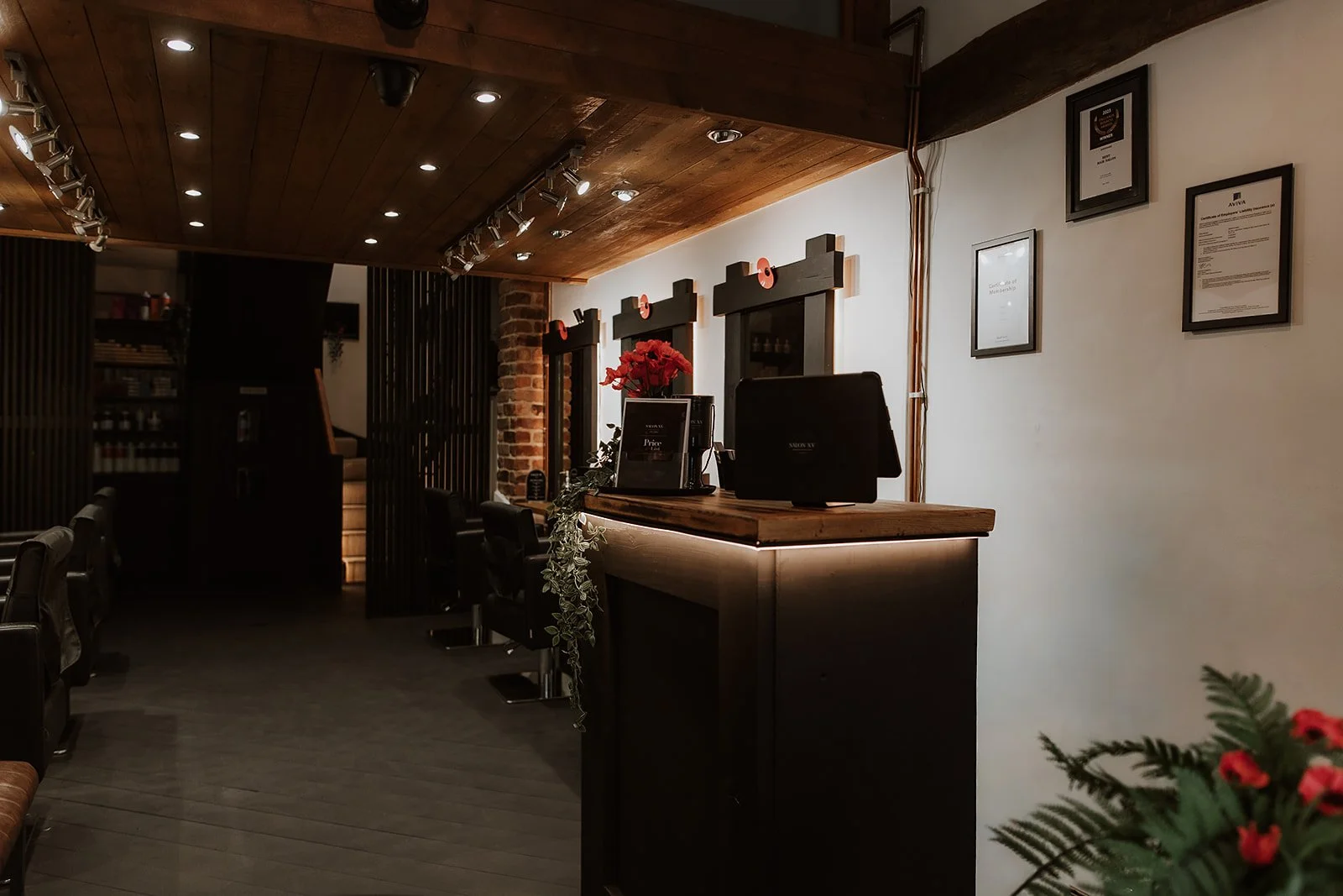 The reception desk of Salon XV in Oswestry with computer monitors, framed certificates on the wall, hanging flower arrangements, and warm wooden accents on the ceiling and counter.