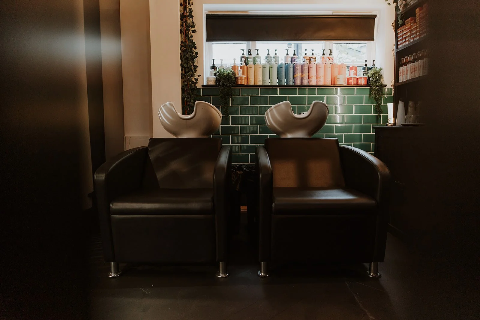 Two black salon chairs with white ceramic hair wash basins behind them, placed in front of a green tiled wall. Above the window, there are various colourful hair product bottles.