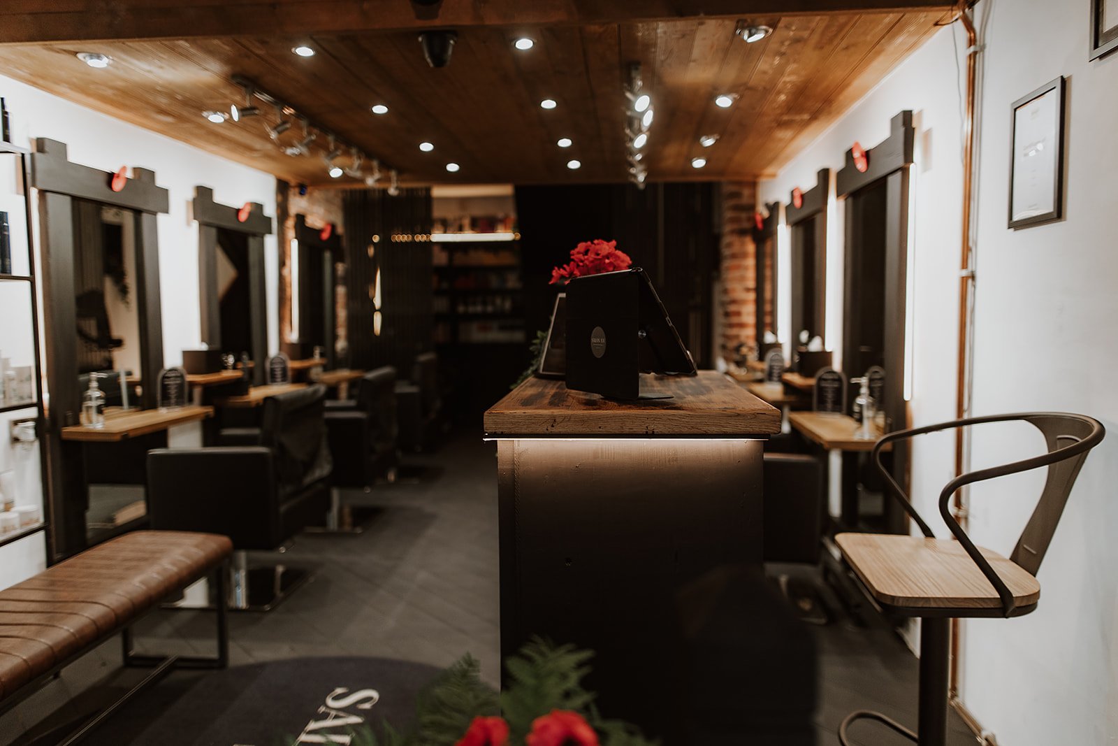 Interior of a modern hair salon with individual styling stations along the wall, each with a mirror and salon chair. A wooden ceiling with recessed lights and track lighting, with some decorations and framed pictures on the wall. Salon XV Oswestry.