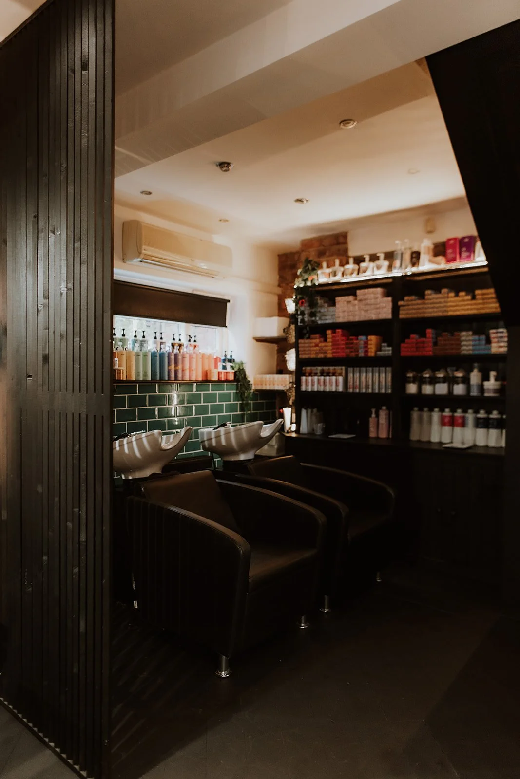 Interior of a Salon XV in Oswestry with black chairs, washing stations, shelves with hair products, and a window with a dark roller shade.