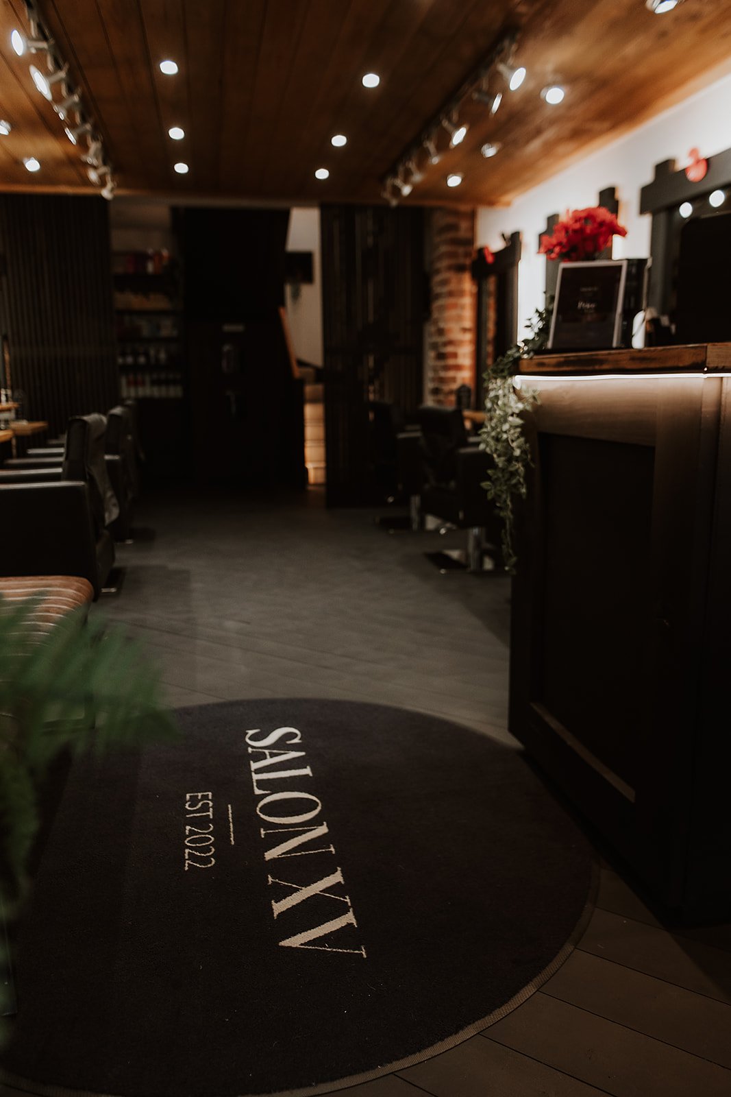 Interior of Salon XV in Oswestry with a wooden ceiling, black chairs, and decorative lighting. There is a black rug with white text reading 'SALON XV EST 2002' at the entrance.