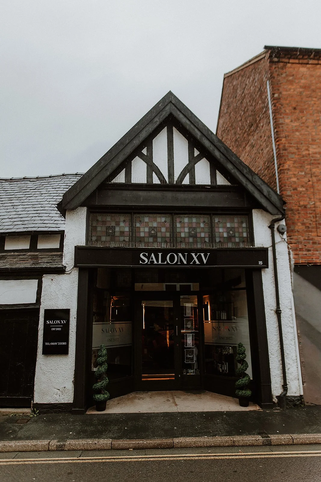 Exterior of a building on Beatrice Street in Oswestry with a sign that reads 'Salon XV,' featuring a black and white Tudor-style facade, glass door entrance, and two small topiary plants at the front.