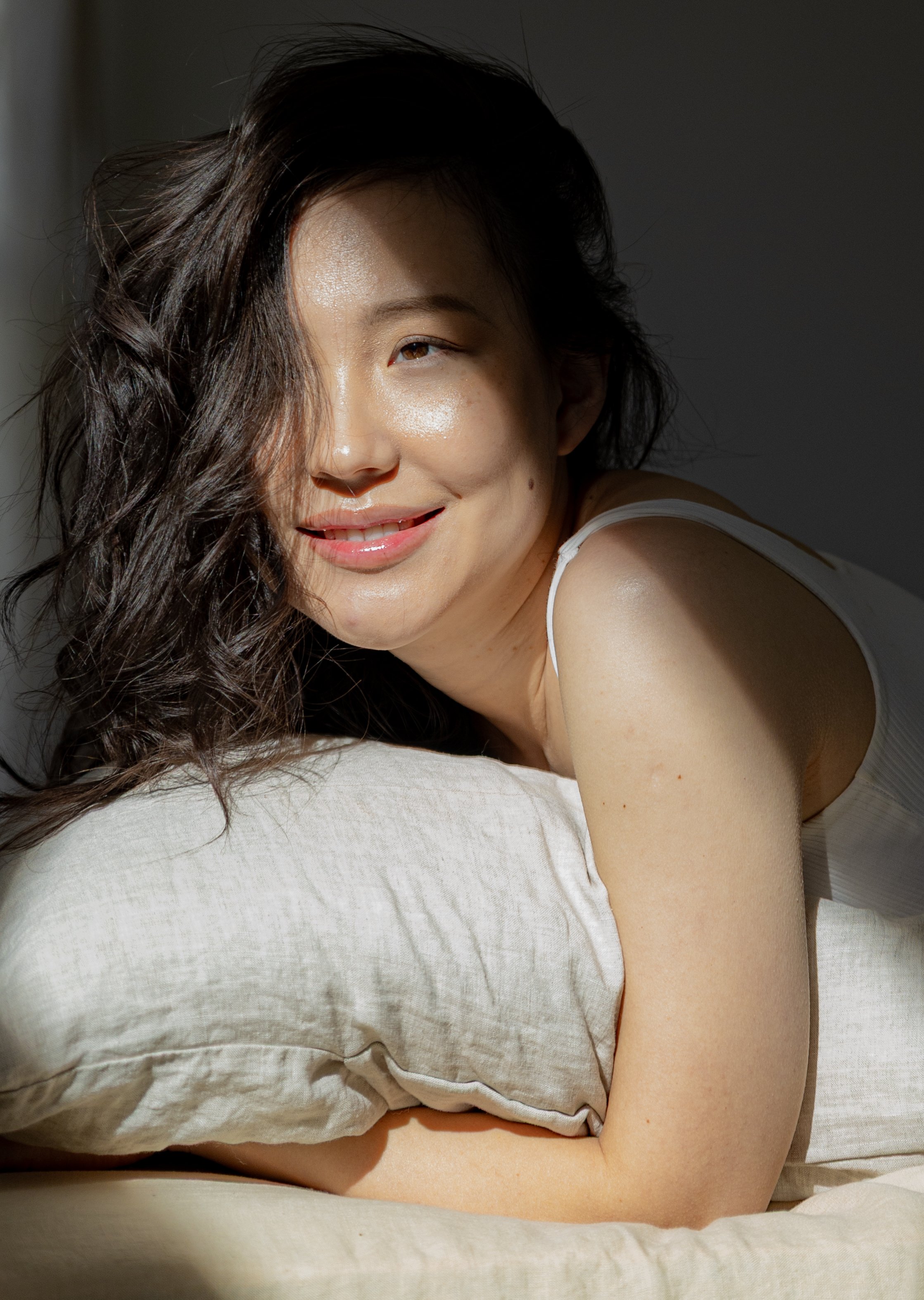 A woman with wavy dark hair lying on a pillow, smiling with her face turned towards the camera in natural light.