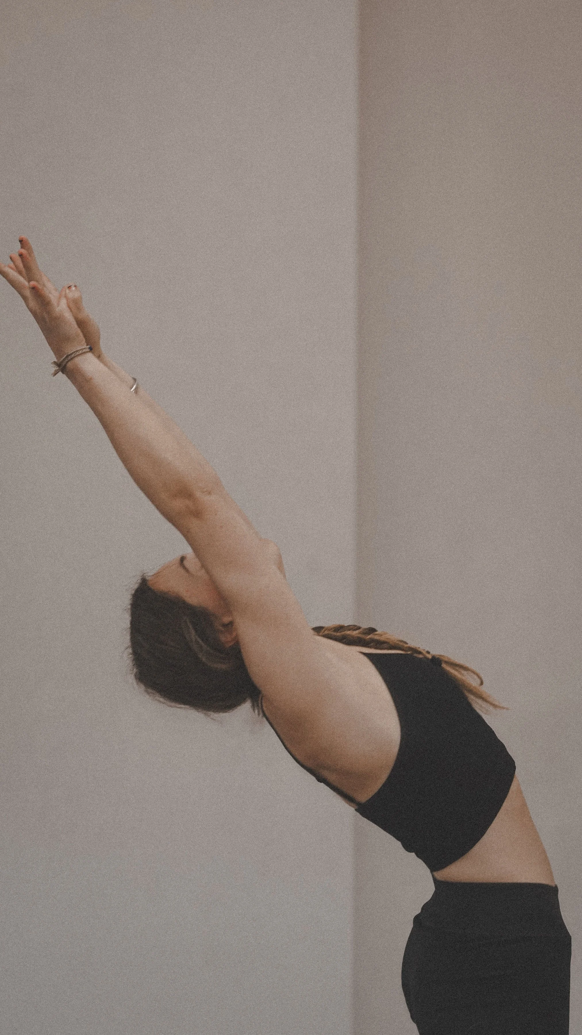 A woman in a black sports bra and pants performing a yoga pose with arms extended overhead and body arched backward.