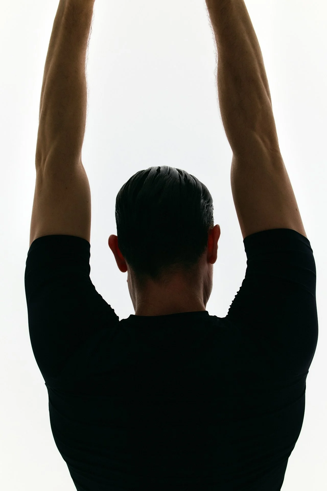 Back view of a man with short dark hair, wearing a black shirt, raising both arms above his head against a plain white background.