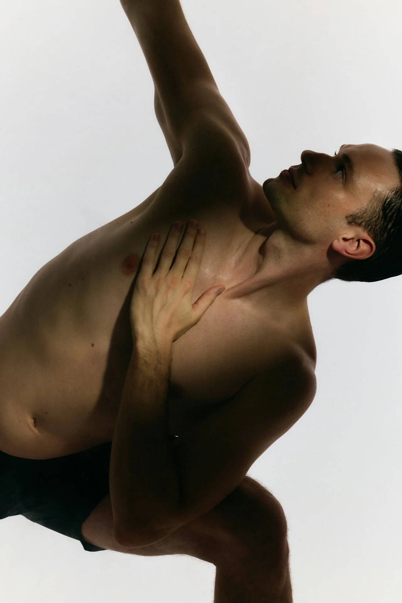 A shirtless young man with short dark hair and light skin, with his head tilted back and looking to the side, raising one arm, against a plain white background.