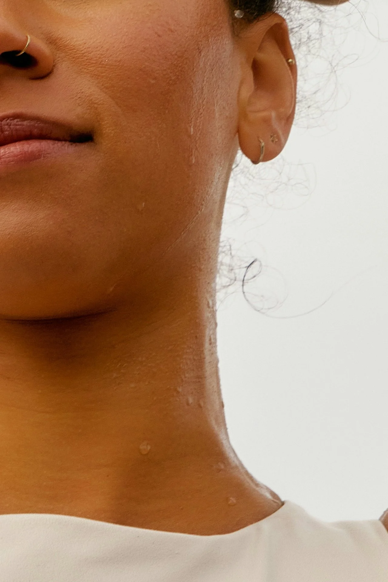 Close-up of a woman's face, neck, and part of shoulder, with water droplets on her skin, wearing small earrings and a white top.