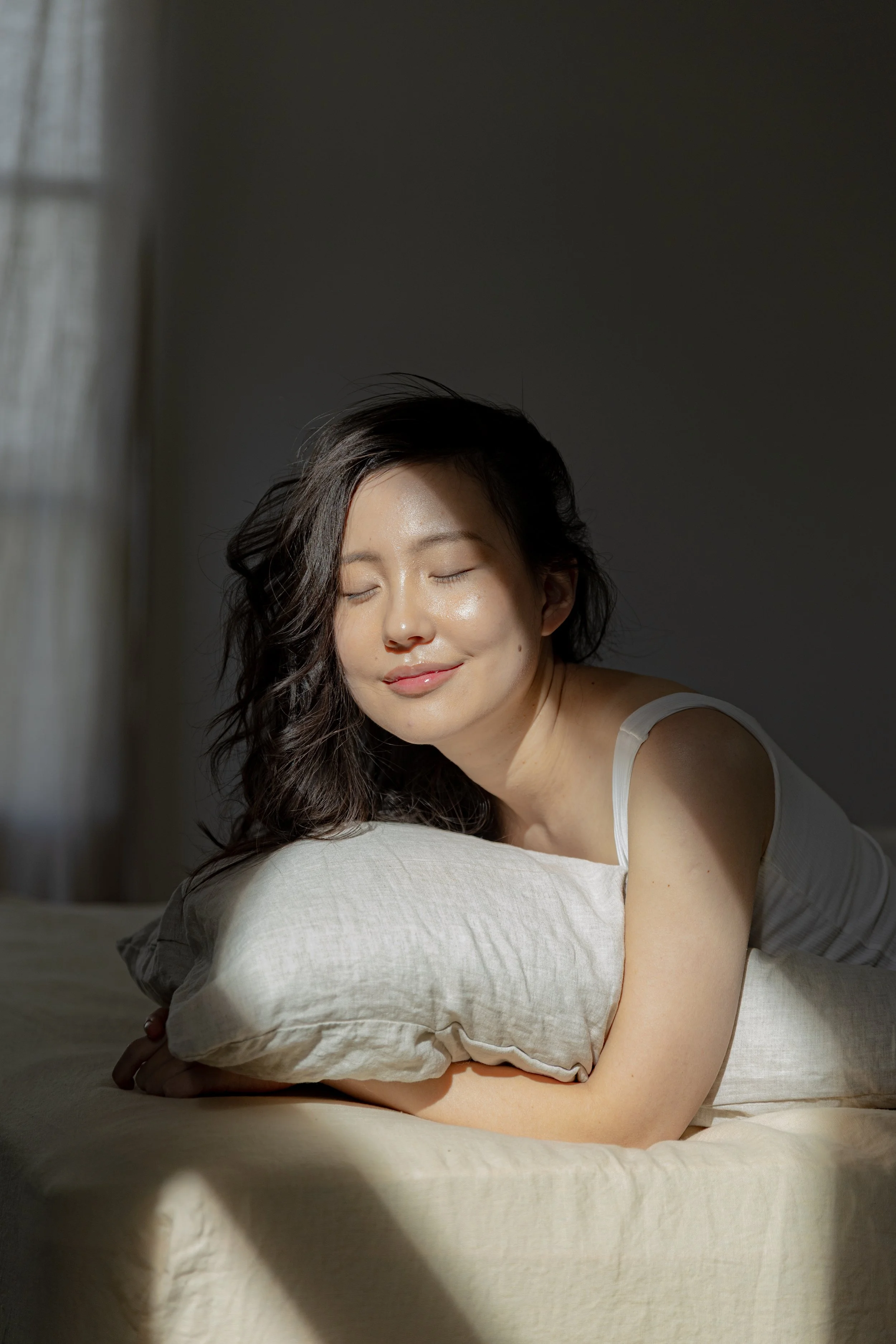 A woman with dark wavy hair smiling with eyes closed, resting her head on a pillow on a bed, with sunlight and shadows in the background.