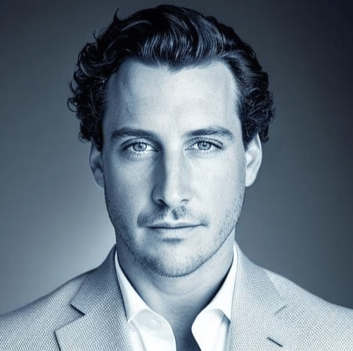 A black-and-white portrait of a young man with wavy dark hair, wearing a light-colored blazer and white shirt, looking directly at the camera.