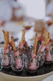 Six shot glasses filled with pink substance, each garnished with a pretzel stick and a piece of ham.