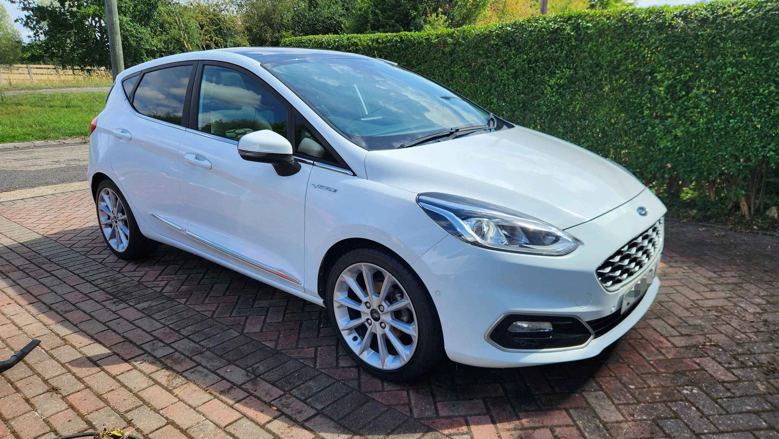 A white hatchback car parked on a brick driveway next to a hedge.