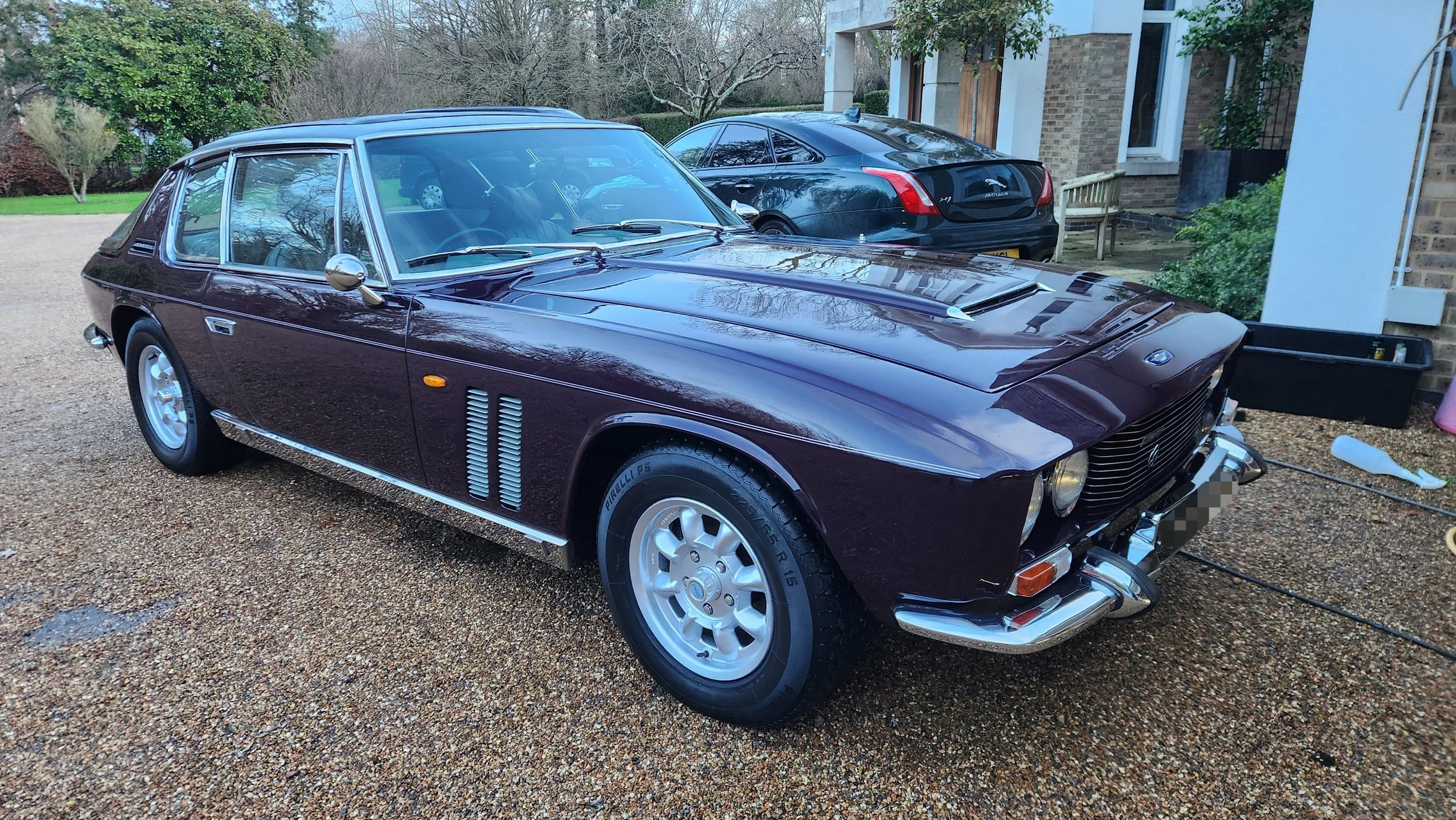 A vintage maroon car parked on a gravel driveway beside a black modern car, with a house and greenery in the background.
