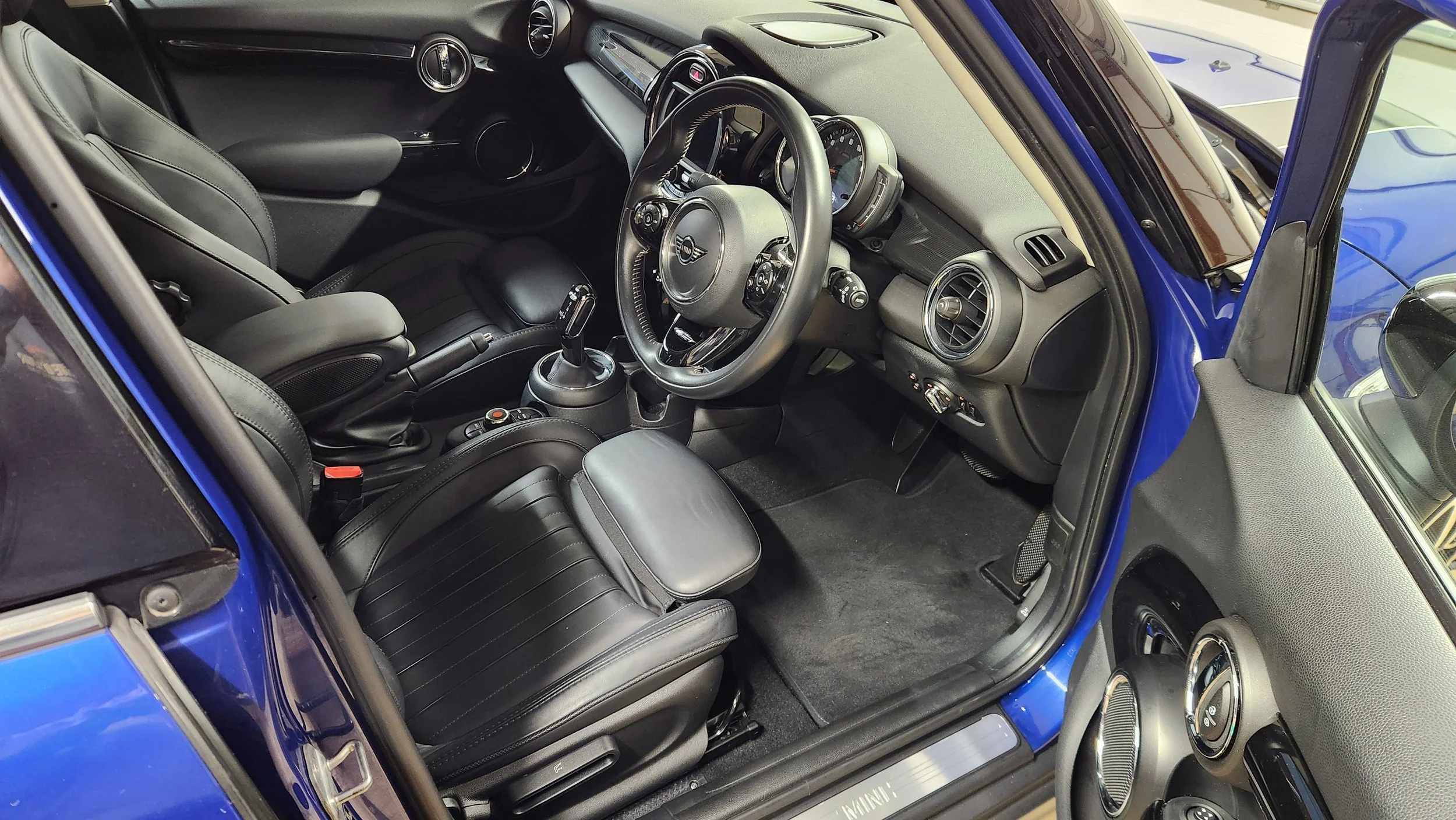 Interior of a blue MINI Cooper, showing black leather seats, steering wheel with MINI logo, dashboard with circular air vents, gear shift, and audio controls.