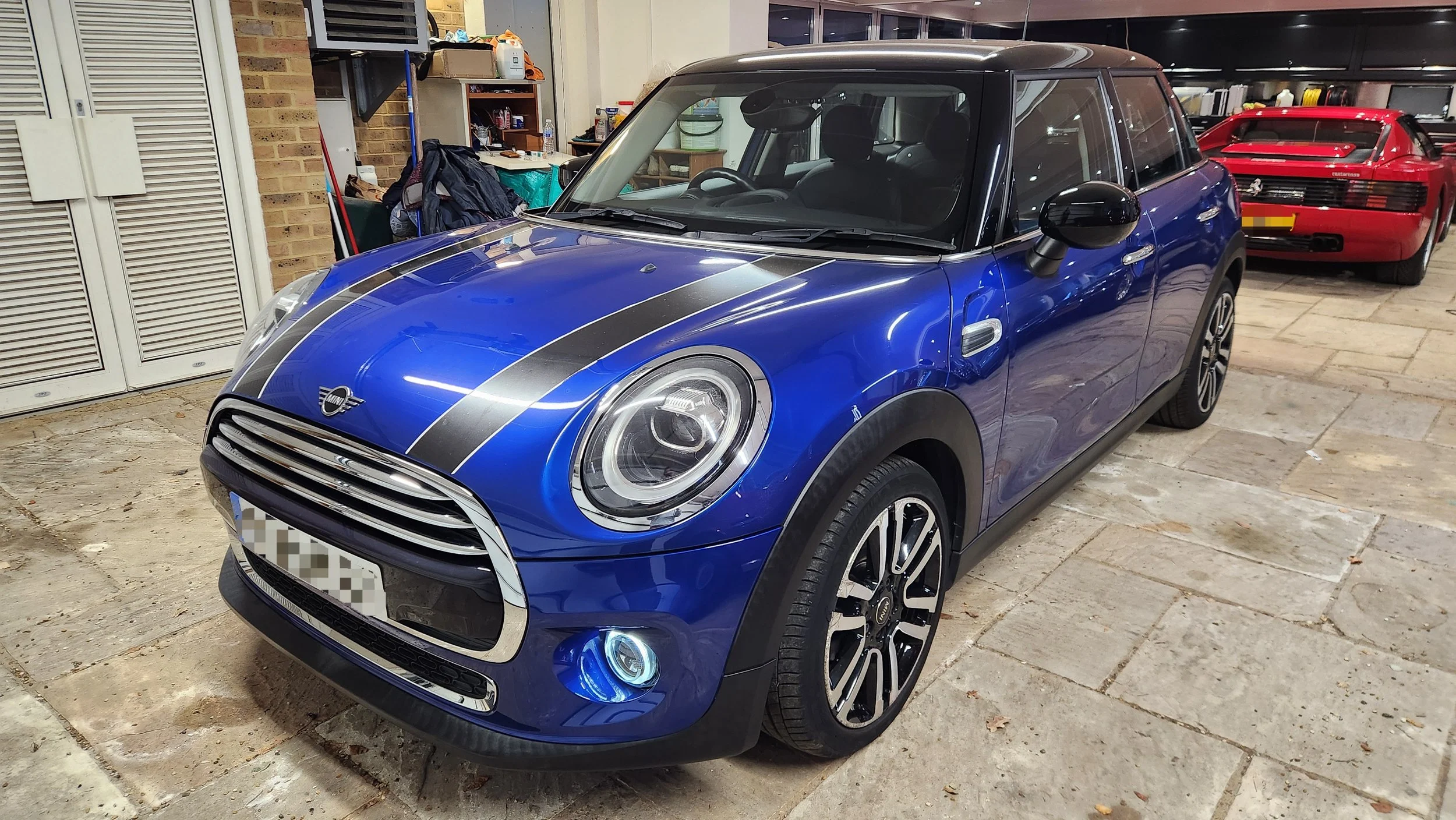 Blue Mini Cooper with black racing stripes parked in garage with another red sports car in background.