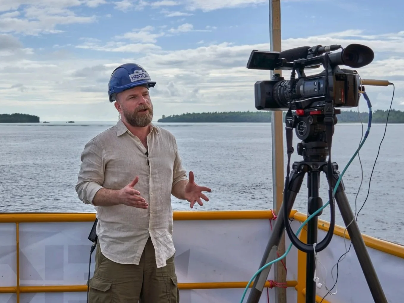 A man with a beard wearing a white shirt and a blue helmet standing next to a video camera in front of the sea